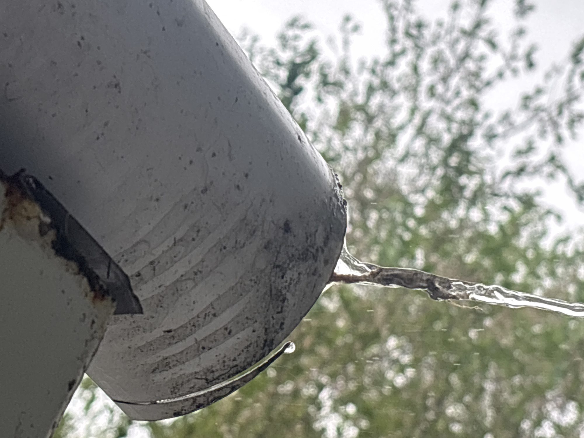 A close-up, low-angle shot captures a weathered, grey downspout or gutter exit with water flowing out. The spout, likely made of metal, exhibits signs of age with rust-colored patches and grime along its edges and interior. Horizontal striations are visible on the inner surface of the spout, possibly from its construction or wear.
A clear stream of water extends horizontally from the spout's opening, appearing vibrant against the subdued tones of the metal. A single, distinct water droplet clings to the underside of the spout's lip, reflecting the light.
The background is softly blurred, dominated by out-of-focus green foliage, likely trees or bushes, set against a bright, possibly overcast sky. This soft bokeh effect emphasizes the foreground elements. The presence of flowing water suggests either ongoing rainfall or recent precipitation. No people or text are visible in the image. The scene conveys an outdoor, potentially rainy, environment.