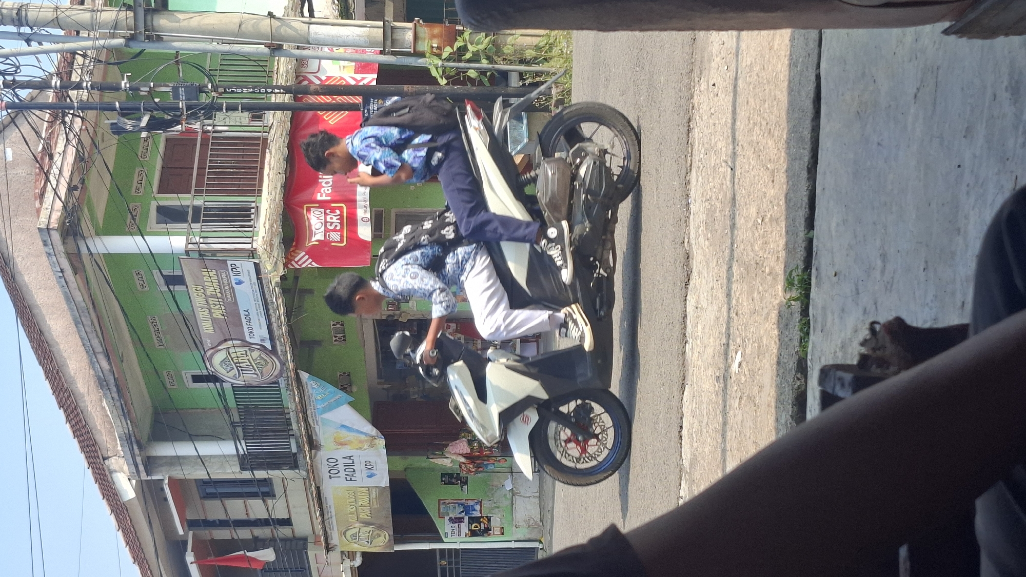 Two Indonesian male students, wearing school uniforms consisting of blue and white patterned shirts and dark pants, ride on a white scooter. The student in front is driving, while the student behind him is a passenger. Both students have backpacks. The scooter is traveling on a road in front of buildings. The buildings have green facades and balconies. There are banners on the buildings. One banner displays the text "JUARA" within a circular logo, and another reads "KUALITAS TINGGI PASTI JUARA!". A red banner with the text "TOKO SRC Fadi" is also visible. The scene appears to be in an urban or semi-urban area, possibly a commercial street in Cilaku, Indonesia, given the presence of shops and street signage. The lighting suggests it is daytime with sunlight casting shadows.