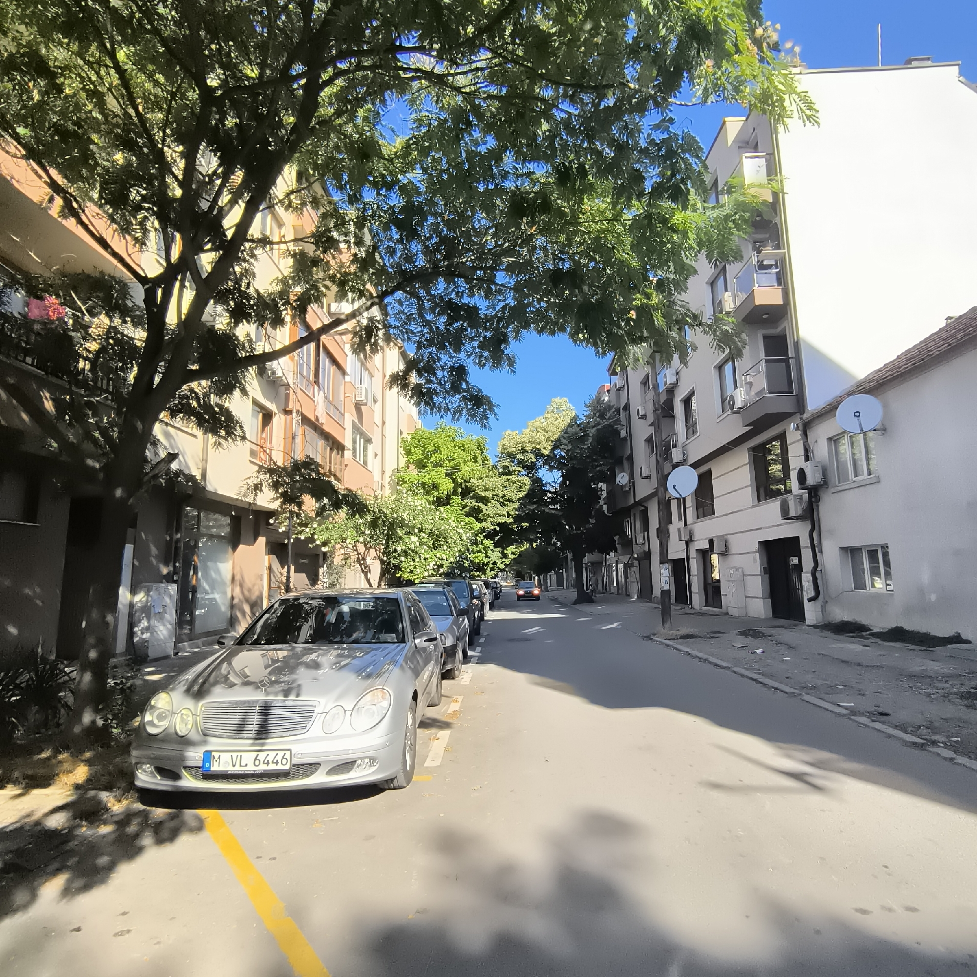 The image depicts a sun-drenched street scene in the city of Varna, Bulgaria. The perspective is at street level, looking down a relatively narrow residential street lined with multi-story buildings on both sides. The foreground is dominated by a silver Mercedes-Benz sedan, parked parallel to the curb and partially obscured by the dappled shade of a large, leafy tree. A bright yellow painted line, indicative of parking regulations, is visible on the asphalt in front of the car.
The buildings on the left appear to be apartment blocks, characterized by varying facade colors and window arrangements. On the right, the buildings are a mix of styles, with one appearing to be a lighter-colored apartment building and another with a more traditional tiled roof. Several satellite dishes are mounted on the exterior walls of the buildings. The street itself is paved and appears to be a two-way road with parked cars lining the left side. In the distance, more trees and buildings are visible, and a single car with its headlights on is moving away from the camera. The sky is a clear, bright blue. The overall impression is of a quiet, typical urban residential area on a sunny day. There are no people visible in the scene.