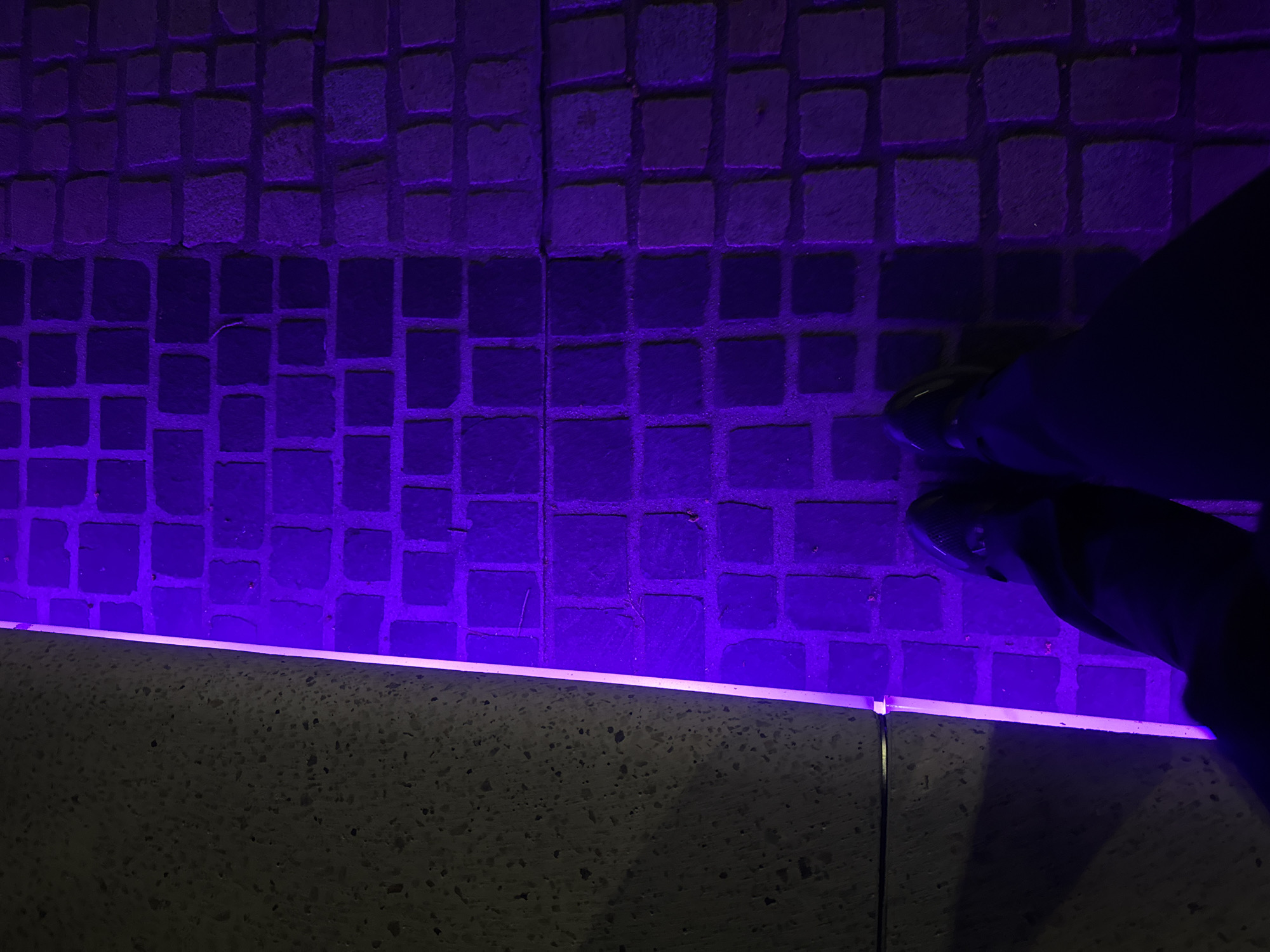 An overhead view captures a nighttime or artificially lit scene in Perth, Australia, dominated by vibrant purple illumination. The ground consists of dark, textured, square-to-rectangular paving stones, resembling cobblestones, which are brightly lit by a linear purple light strip running horizontally across the bottom third of the frame. This light strip is integrated into the top edge of a darker, textured concrete or stone wall or ledge. On the right side of the image, the lower legs and feet of a person are visible from an aerial perspective. The person wears dark trousers and dark shoes with a slight sheen or metallic detail, casting a faint shadow on the purple-lit paving. The overall atmosphere is modern and urban, created by the interplay of the patterned ground, architectural lighting, and the human presence. There is no visible text.