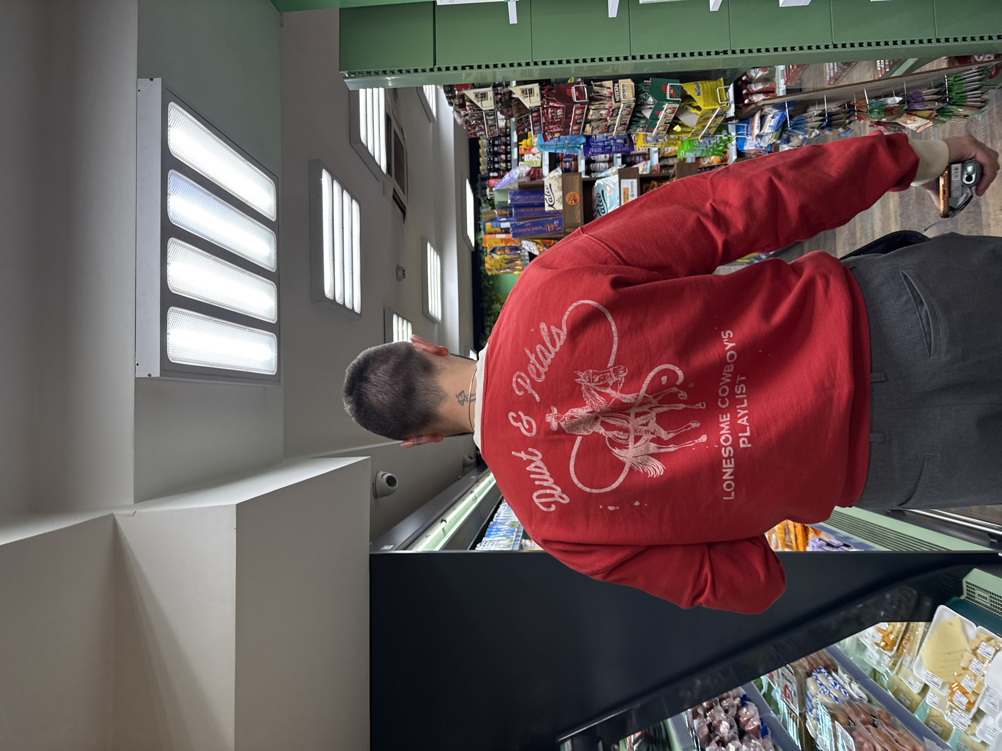 A man wearing a red sweater with a white print of a horse and rider and the words "Dust & Petals" and "Lonesome Cowboys Playlist" is standing in a store, possibly in Yerevan, Armenia. He is holding a cell phone in his right hand, possibly taking a picture. The store has shelves stocked with various products, including food items. Overhead lighting panels are visible in the ceiling. The man is wearing gray pants.