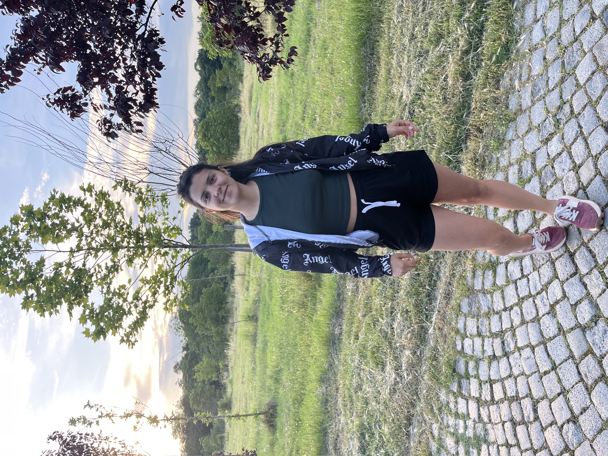 A young woman stands upright on a grey cobblestone path, facing slightly left and smiling gently at the camera. She has dark hair and is dressed in a dark green crop top, black shorts with a white drawstring, and a black and white jacket featuring the word "Angel" printed multiple times on its sleeves. She wears maroon or pink sneakers with white laces. To her left, a wide expanse of green grass leads to a line of trees in the background. The sky above is bright with scattered clouds, displaying the warm, soft light of late afternoon or early evening, possibly near sunset. Tree branches with both dark red and green leaves are visible in the foreground, framing the outdoor scene. The overall setting appears to be a park or natural recreational area.