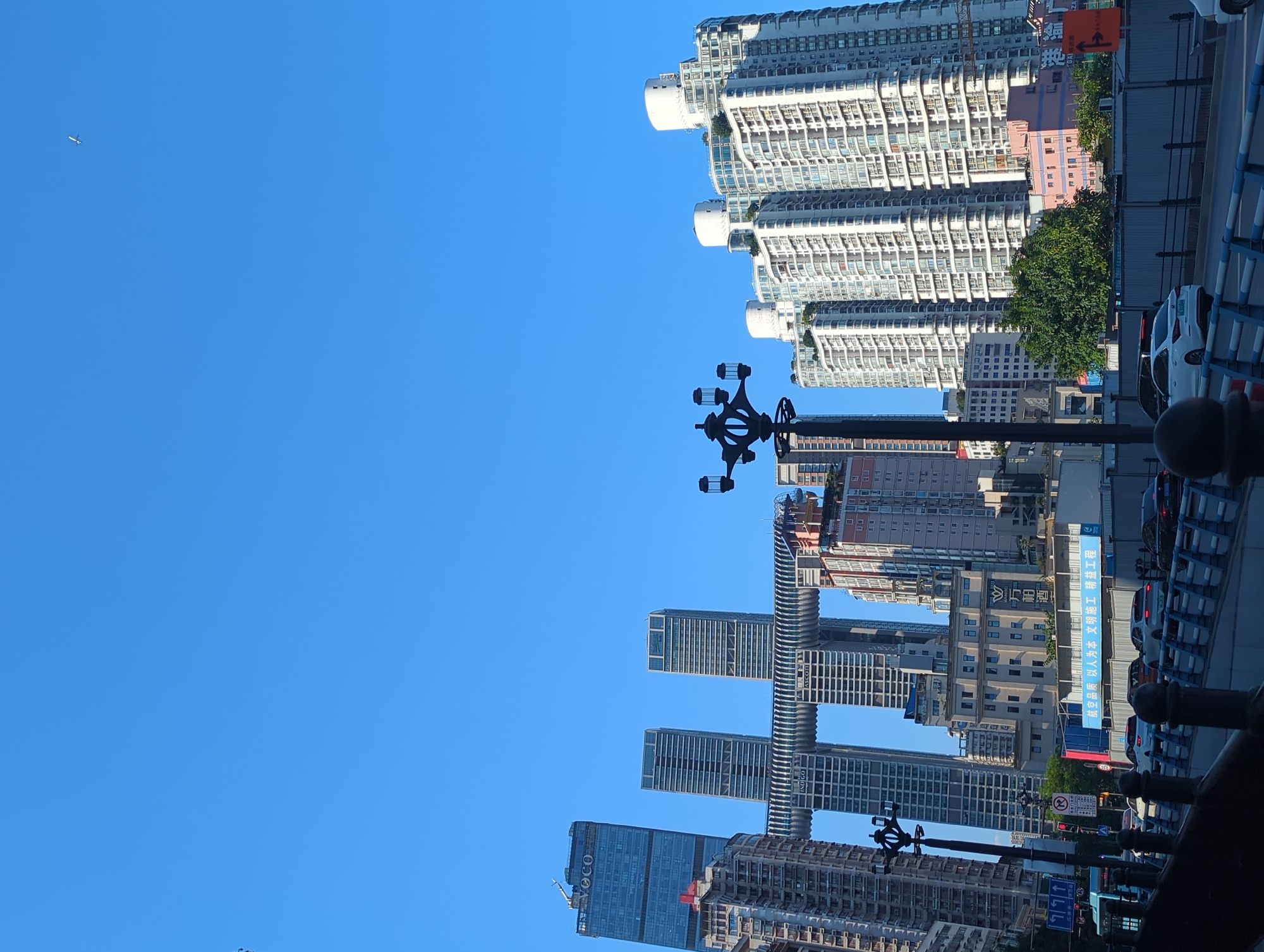 The image captures a cityscape dominated by modern skyscrapers against a clear blue sky. In the foreground, a dark, ornate lamppost stands on the left, partially obscuring a street scene. A blue bus is visible on the left side of the road, and several cars are driving on a bridge or elevated roadway. The road is bordered by a dark railing with blue accents. In the midground, several tall buildings are prominent. One building on the left has "VOCO" signage at its top. Two central buildings are connected by a distinctive horizontal structure with a patterned facade. To the right, a cluster of white, stepped skyscrapers with numerous balconies and windows rises against the sky. The background features more buildings of varying heights and architectural styles. The scene suggests a bustling urban environment, likely in a modern Chinese city. While the exact location is not confirmed from the image alone, the architectural style and signage, such as the Chinese characters on some buildings, point towards a location in China.