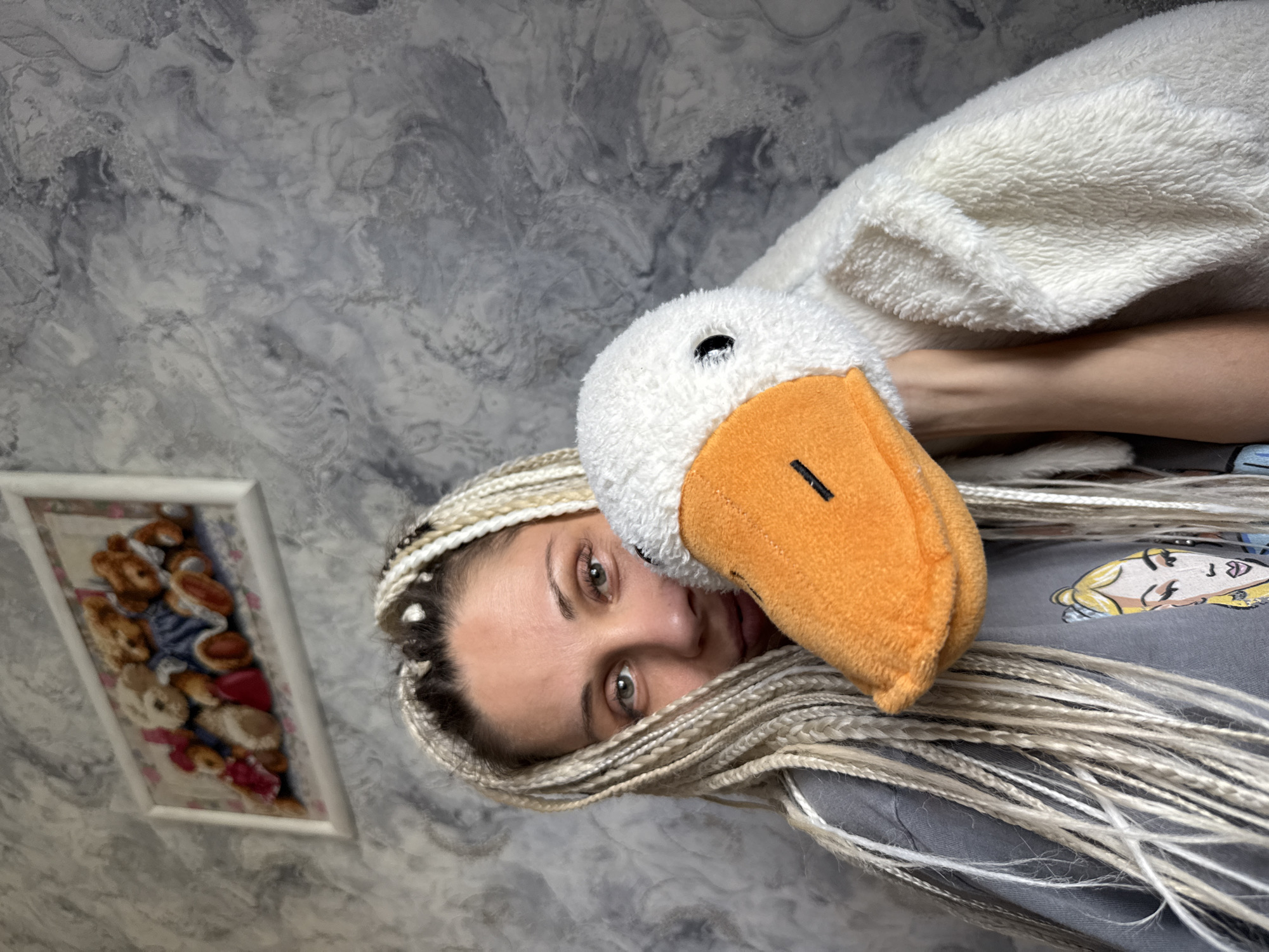 A woman with blonde braids is holding a stuffed goose in front of her face, obscuring the lower half of her face. She is wearing a grey t-shirt with a cartoon image on it. Behind her, a framed picture of teddy bears hangs on a textured grey wall. The image appears to be taken indoors. There is no information available to determine the location in the city of Yaroslavl, Russia.