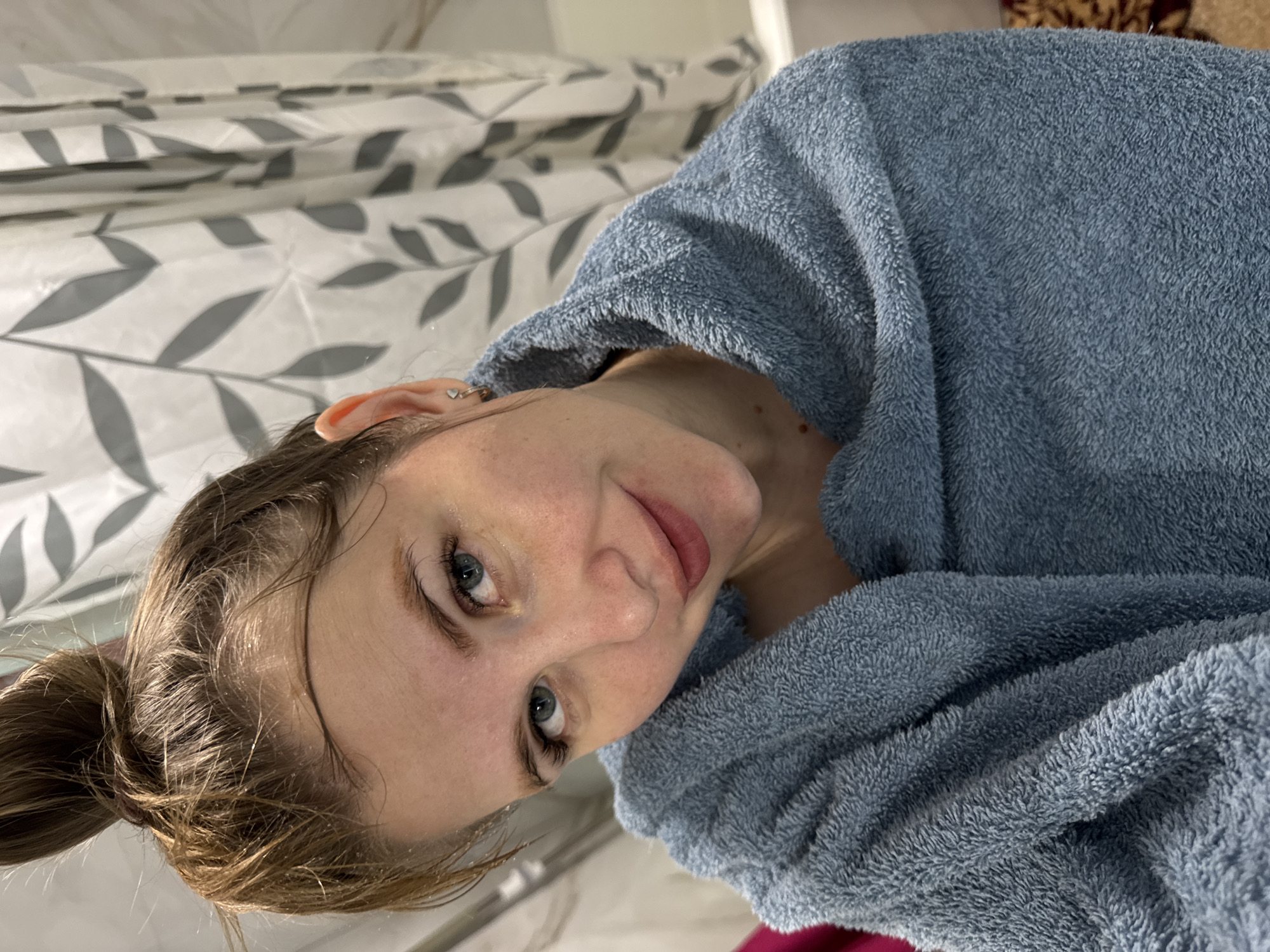 A woman with fair skin and blue eyes is photographed indoors, likely in a bathroom based on the shower curtain in the background. She is wrapped in a blue, plush robe or towel. Her wet hair is pulled back into a bun. She is looking at the camera. The location cannot be determined from the image.