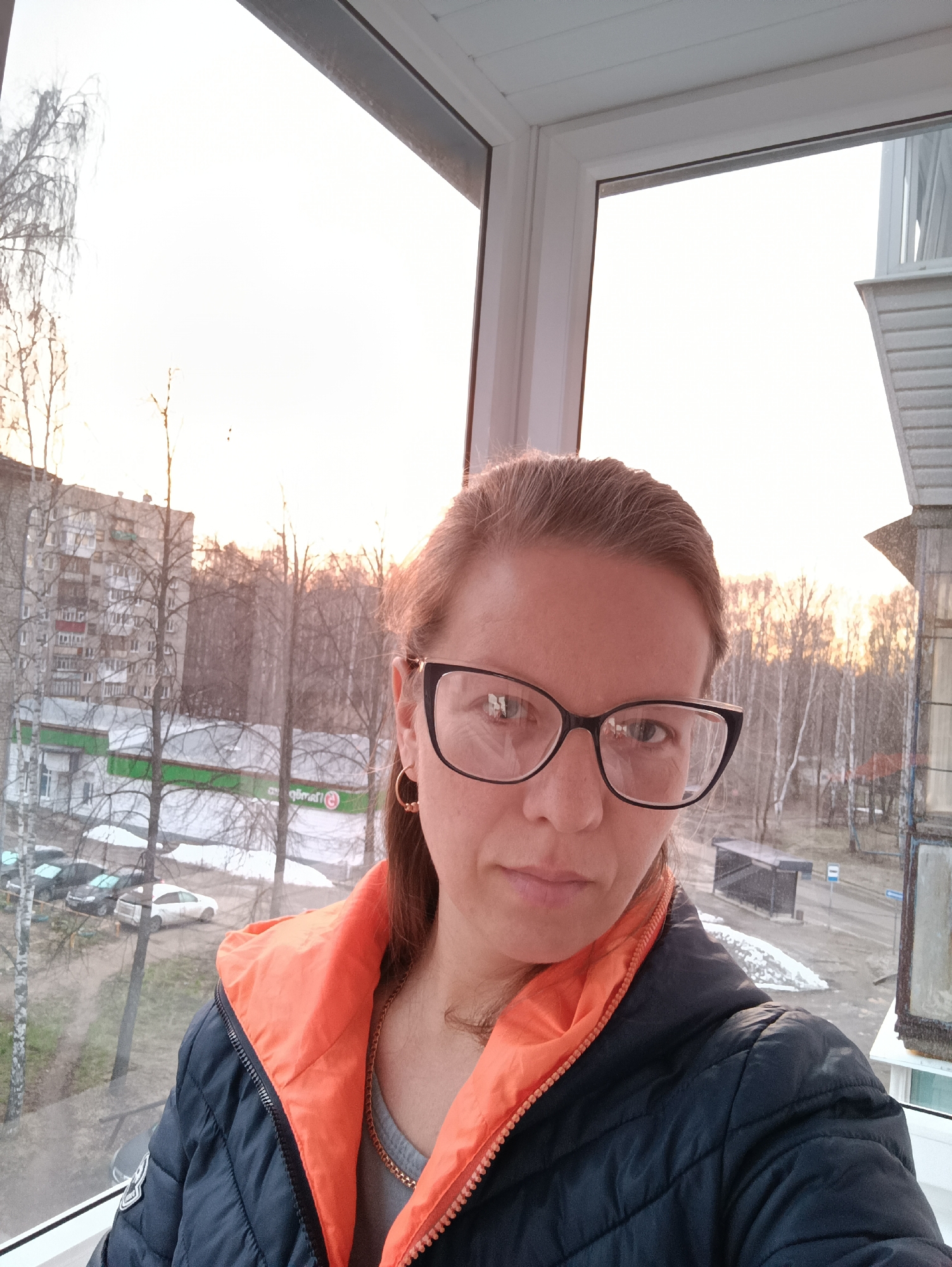A woman is taking a selfie indoors, likely on a balcony or enclosed porch. She is wearing glasses and a dark blue quilted jacket with an orange lining visible at the collar. She has gold hoop earrings. Outside the window are buildings, trees without leaves, and patches of snow. A building with a green roof appears to house a "Магнит" (Magnit) grocery store. The location appears to be Chaikovsky, Russia based on the architecture, store name and Cyrillic lettering. There are a few cars parked near the Magnit building, and a bus stop is visible further down the street. The sky is pale, suggesting either sunrise or sunset.