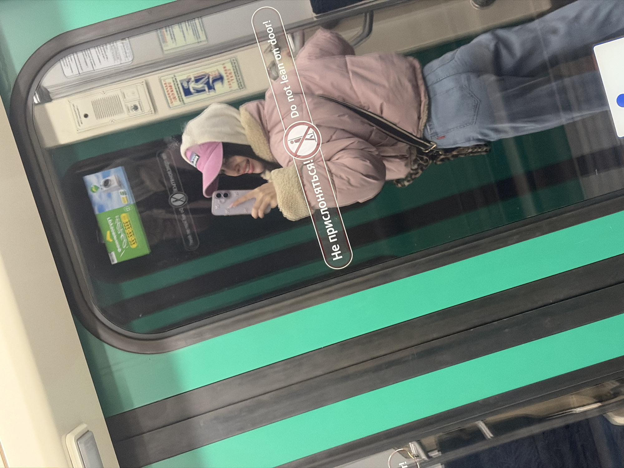 A woman is standing in a train car in Saint Petersburg, Russia. She wears a pink jacket, blue jeans, and a baseball cap with a pink brim. She holds a smartphone in her left hand and looks at the screen. The train car's door is green and black, with stickers and signs. One sign reads, "Do not lean on door!" in English and Russian.