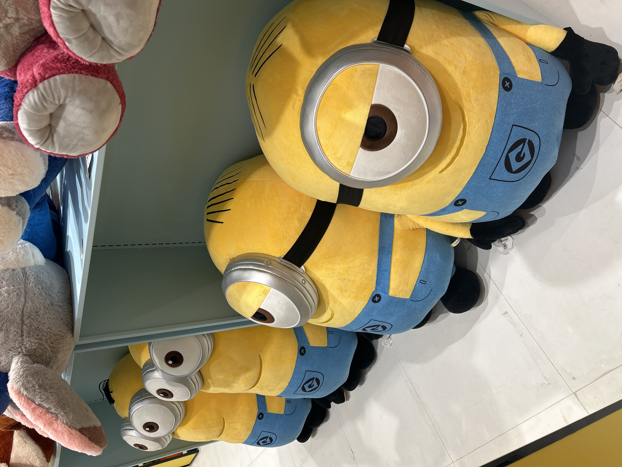 A row of four plush Minion toys, inspired by the animated film franchise, are displayed on a light blue shelf. The Minions are yellow with blue overalls and feature their characteristic single or double eyes with silver goggles. The toys are positioned facing the viewer and are arranged from left to right, with the largest Minion at the front on the right. The shelf is behind the Minions, and above them, other plush toys are visible, including one with pink and grey elements. The floor is tiled with white squares. The scene is brightly lit, suggesting an indoor retail environment. The image does not contain any information that would indicate its location in Makassar, Indonesia.