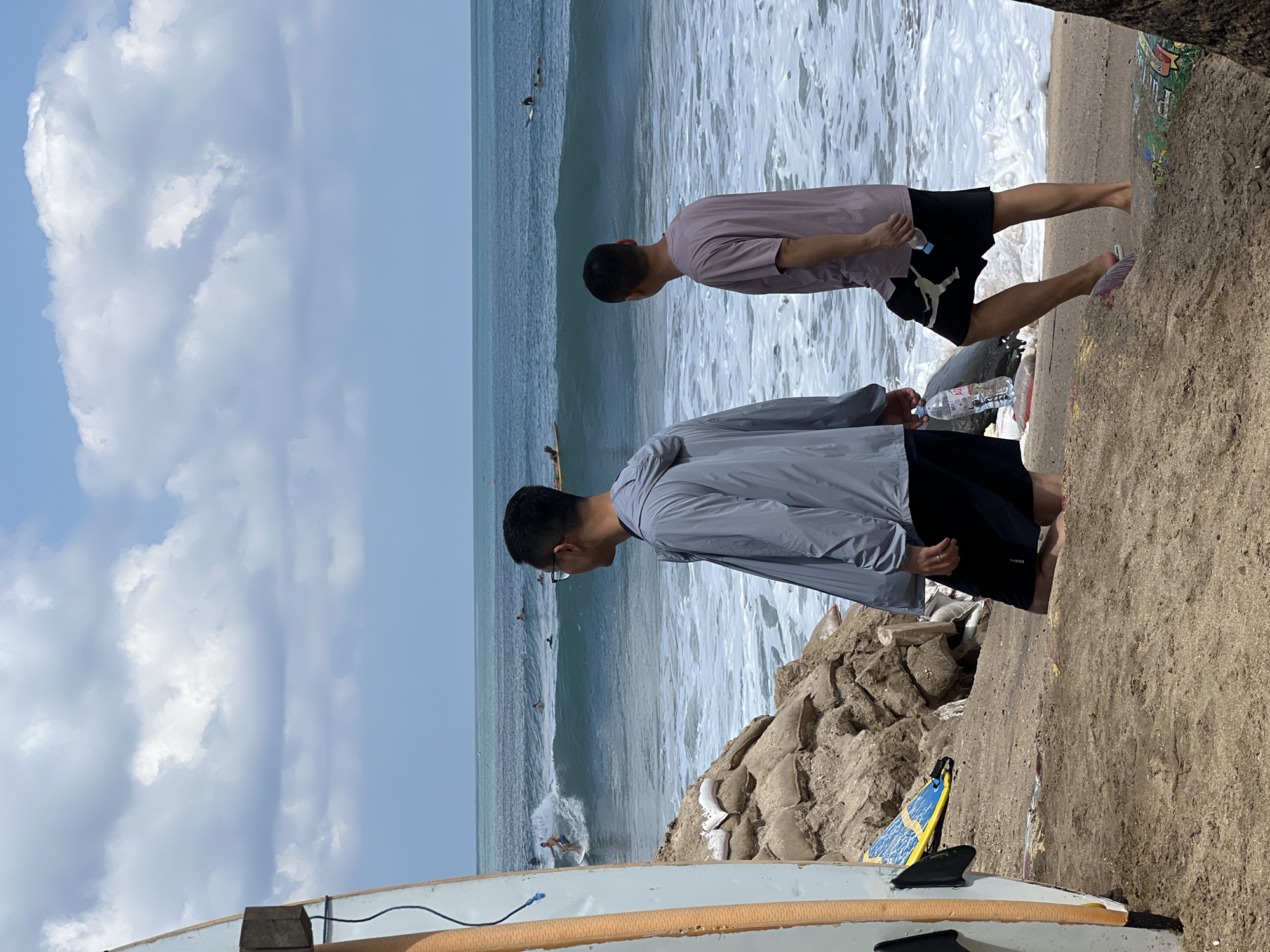 Two men stand on a sandy beach in Kuta, Indonesia, facing the ocean. The man on the left wears a grey hooded jacket and dark shorts, holding a bottle of water. The man on the right wears a light purple t-shirt and black shorts with a white logo, also holding a bottle of water. In the background, the ocean has moderate waves with several surfers visible on the water. A surfboard stands upright on the left side of the foreground. Piles of sandbags are stacked behind the men, likely as a measure against coastal erosion. The sky is partly cloudy.