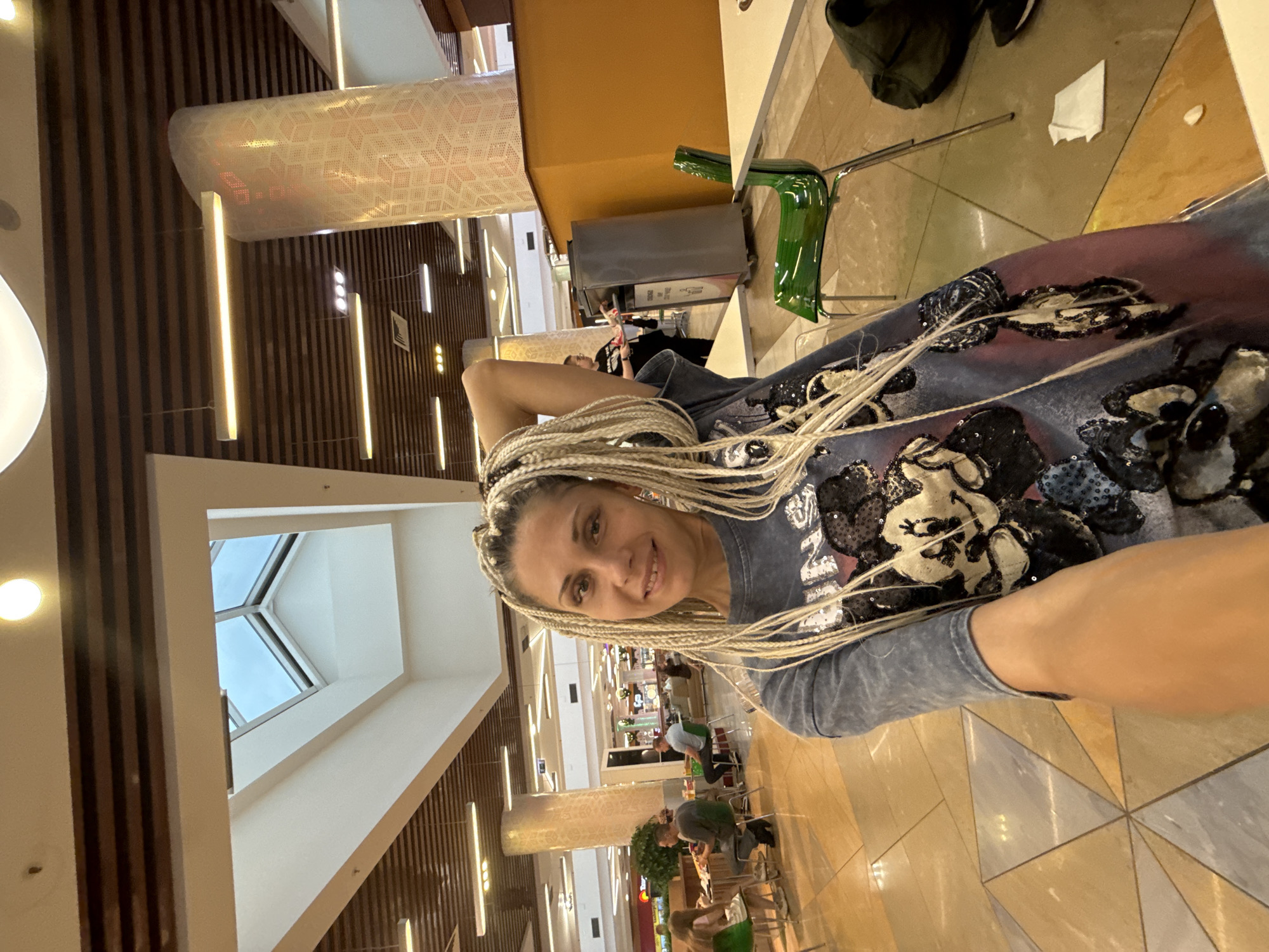 A woman with blonde braided hair is taking a selfie in a brightly lit indoor food court. She is wearing a grey t-shirt with a pattern of cartoon characters. In the background, there are several tables and chairs, as well as other people eating. There is a large pillar with a decorative pattern and a skylight in the ceiling. The floor is tiled in a geometric pattern. The location is not specified as Arkhangelsk, Russia, but appears to be a commercial establishment.