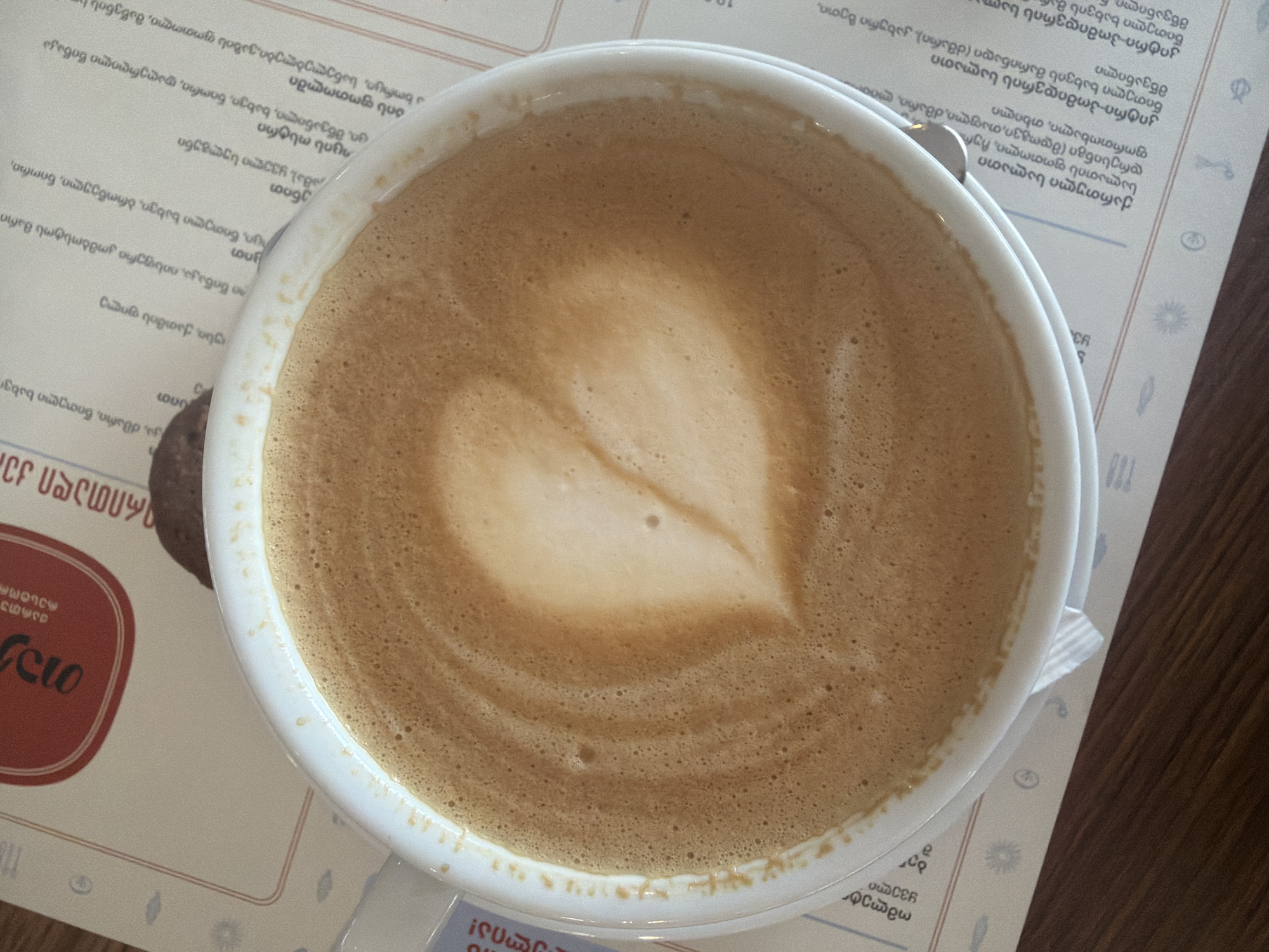 The image shows a top-down view of a cup of coffee, possibly a latte or cappuccino, with latte art in the shape of a heart. The coffee is in a white cup, which is sitting on a paper placemat with text in what appears to be Georgian script and small decorative icons. The surface under the placemat appears to be a wooden table. Part of a red logo is visible on the left side of the image. Although the OCR includes Zeda Bashi, Georgia, the image itself doesn't give specific location information.