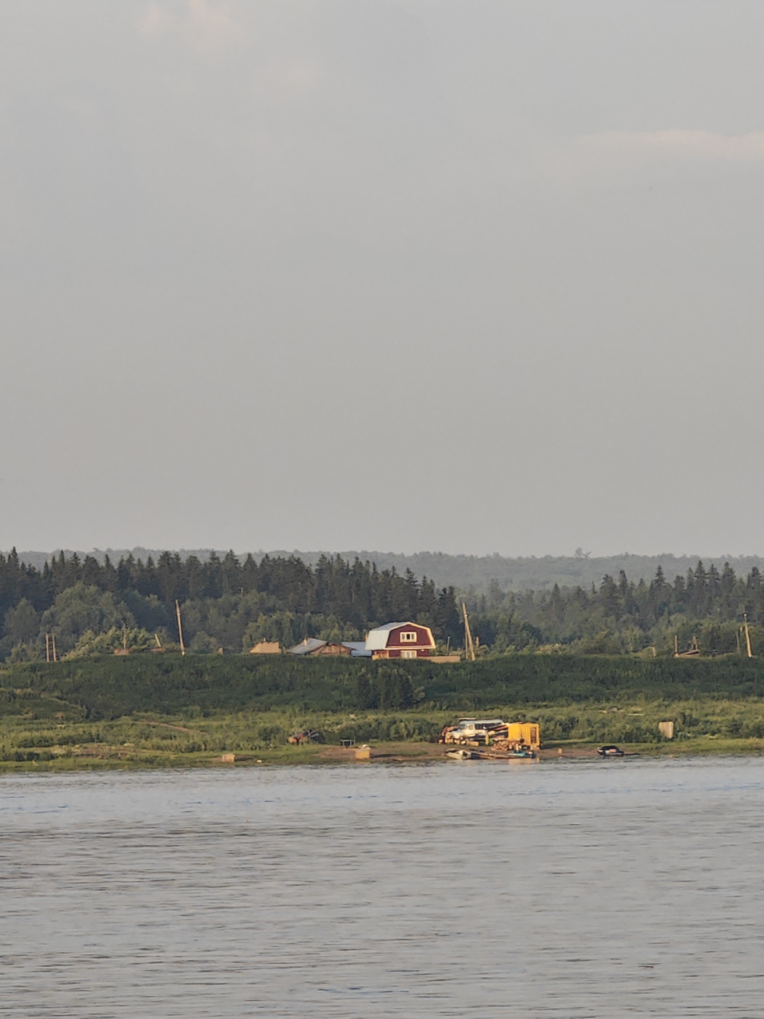 The image shows a wide river in the foreground, reflecting the overcast sky. On the far bank, a verdant slope is covered with grass and vegetation, dotted with trees. A cluster of buildings is situated atop the slope, including a prominent red barn-like structure with white trim and a small window. Further along the bank, several other houses and structures are visible, some with peaked roofs. Behind these buildings, a dense forest of dark green trees stretches across the horizon. On the riverbank in the middle ground, several objects are present, including what appears to be a yellow modular structure, a white vehicle, and a dark-colored car, as well as a small boat. The overall scene suggests a rural or semi-rural setting, possibly a settlement along a river. The location is identified as Lesosibirsk, Russia.