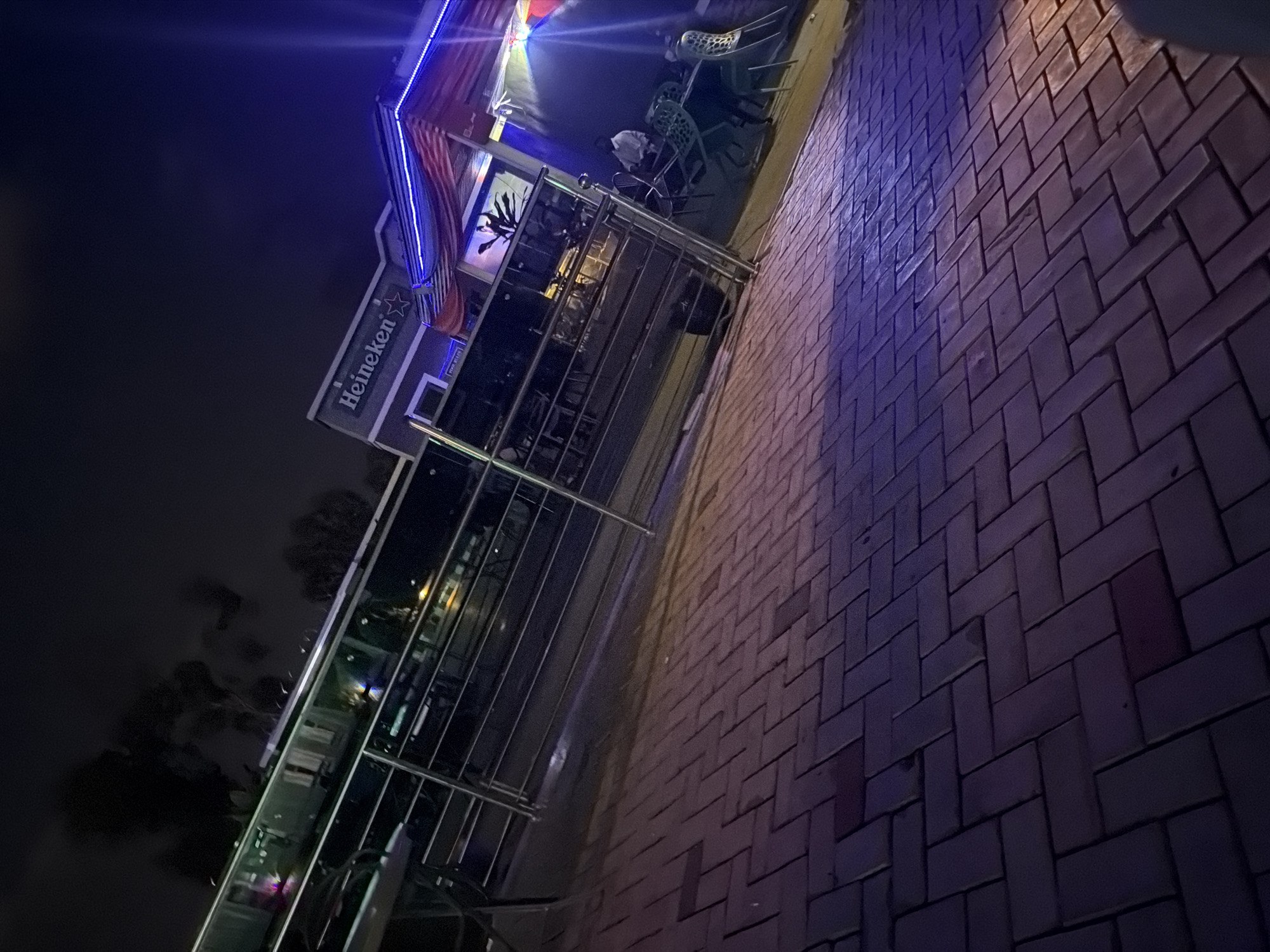 The image depicts an outdoor scene at night, possibly a bar or restaurant, in Bukuru, Nigeria. A prominent sign for "Heineken" is visible on the building. The foreground is dominated by a brick-patterned paved area. In the middle ground, there's a metallic railing and tables and chairs, suggesting an outdoor seating area. A person in a white shirt is seated at one of the tables. The background shows a building with lights and decorative elements, including red and blue lighting. The overall ambiance is illuminated by artificial lighting.