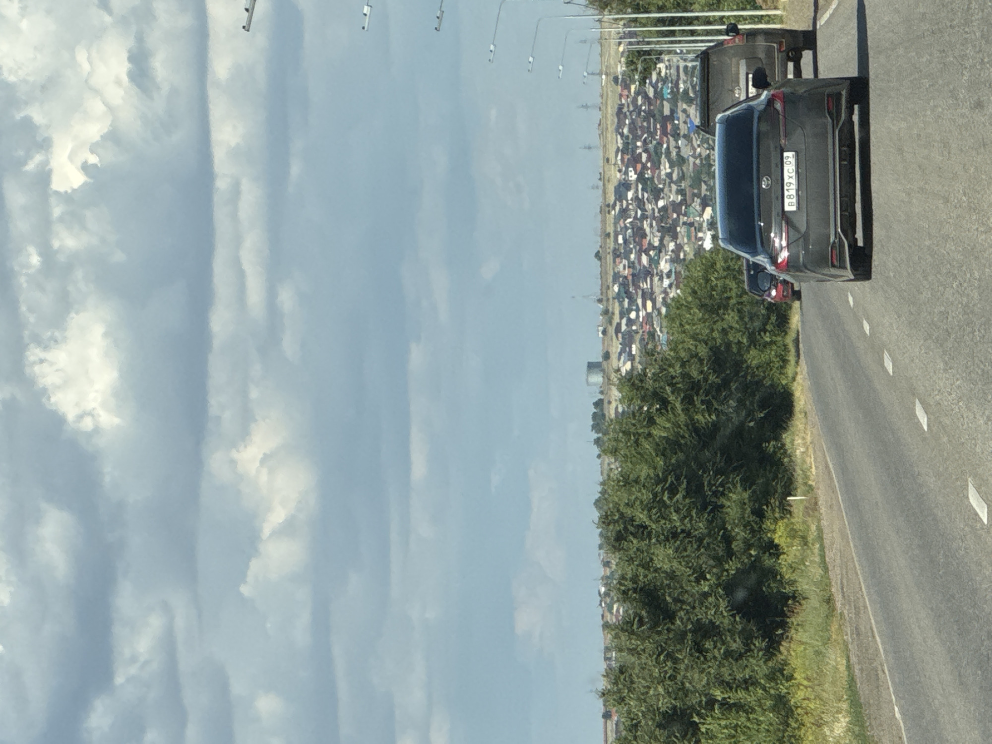 This is a landscape photograph taken from a vehicle moving on a road, oriented vertically. The foreground shows the road surface with white lane markings. To the left of the road is a dense green tree line, and beyond that, a vast expanse of sky filled with scattered white clouds.
In the background, on the right side of the road, a long line of buildings stretches into the distance. The buildings appear to be mostly residential, with a variety of roof colors, creating a dense urban or suburban landscape. Several cars are visible on the road, including a dark-colored SUV in the middle ground, displaying a license plate that reads "B819XC 09". Further ahead, a very long queue of vehicles is visible, suggesting a traffic jam or a procession of some kind. Beyond the buildings, the horizon is hazy.
The scene suggests daytime, with bright sunlight illuminating the clouds and the road. The weather appears to be clear and sunny. The presence of the license plate with Cyrillic characters indicates the location is likely Russia. The overall impression is a snapshot of a journey through a populated area on a sunny day.