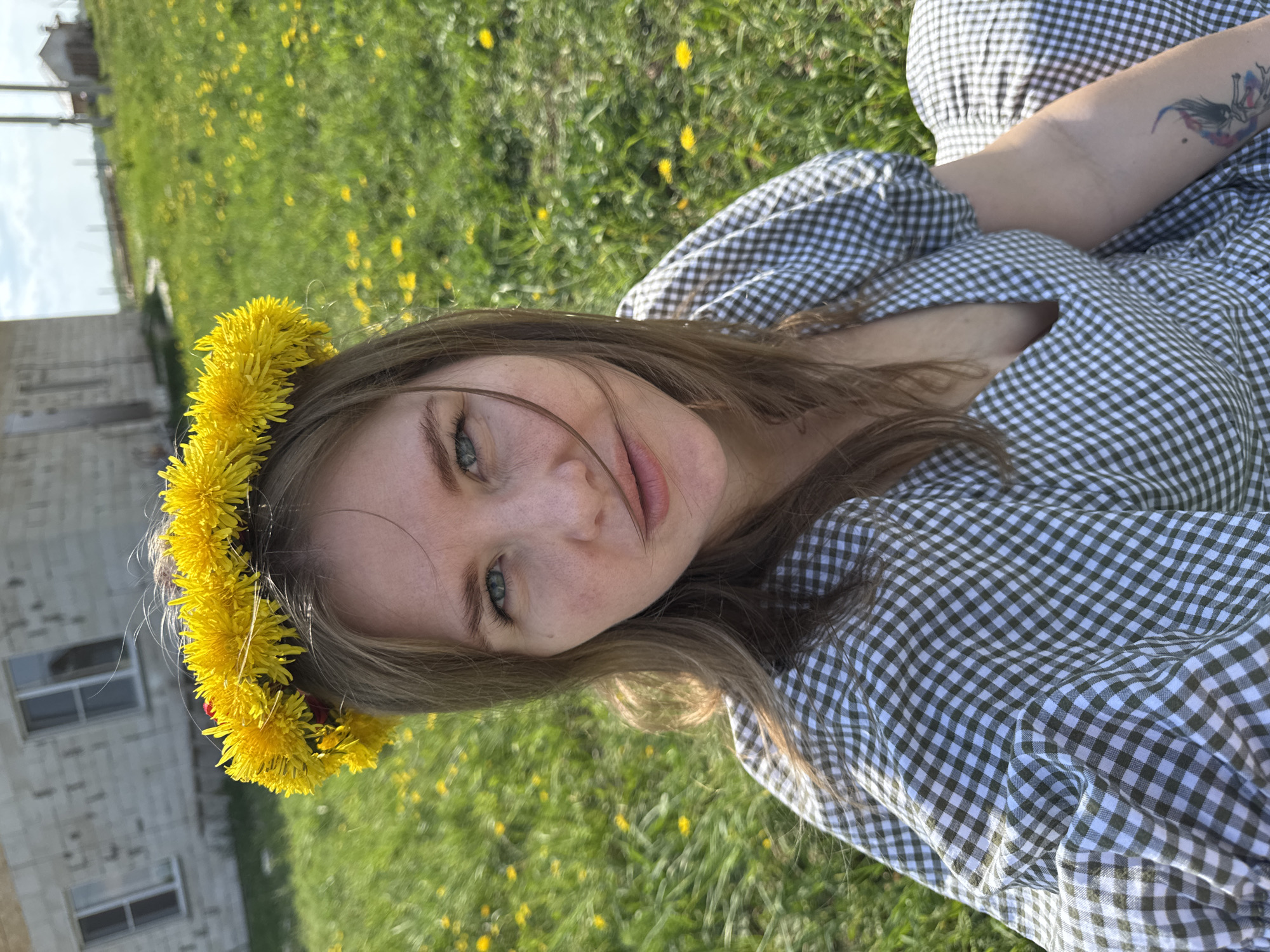 A woman is lying in a grassy field covered in dandelions. She is wearing a green and white gingham dress and a crown of dandelions. Her right arm is visible and has a tattoo. There is a building in the background. The photograph was taken in Ферма, Russia.