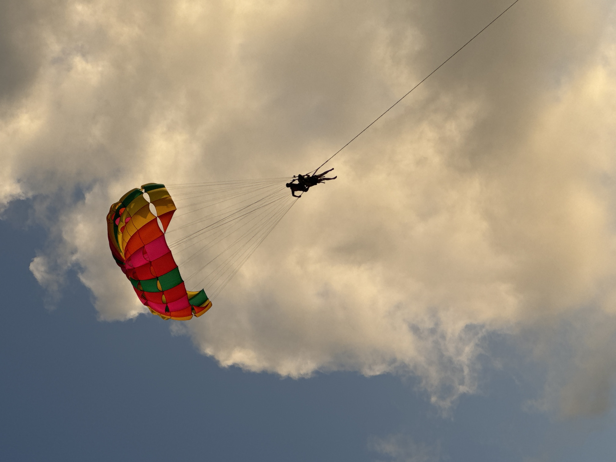 A wide shot captures two individuals parasailing against a dramatic sky over Patong, Thailand. The parasail is vibrant with red, pink, green, and yellow panels, billowing above. Silhouetted against a backdrop of cloudy, golden-hued sky, the two people are suspended mid-air, connected by ropes to the parasail. The lower half of the sky transitions to a cooler blue.