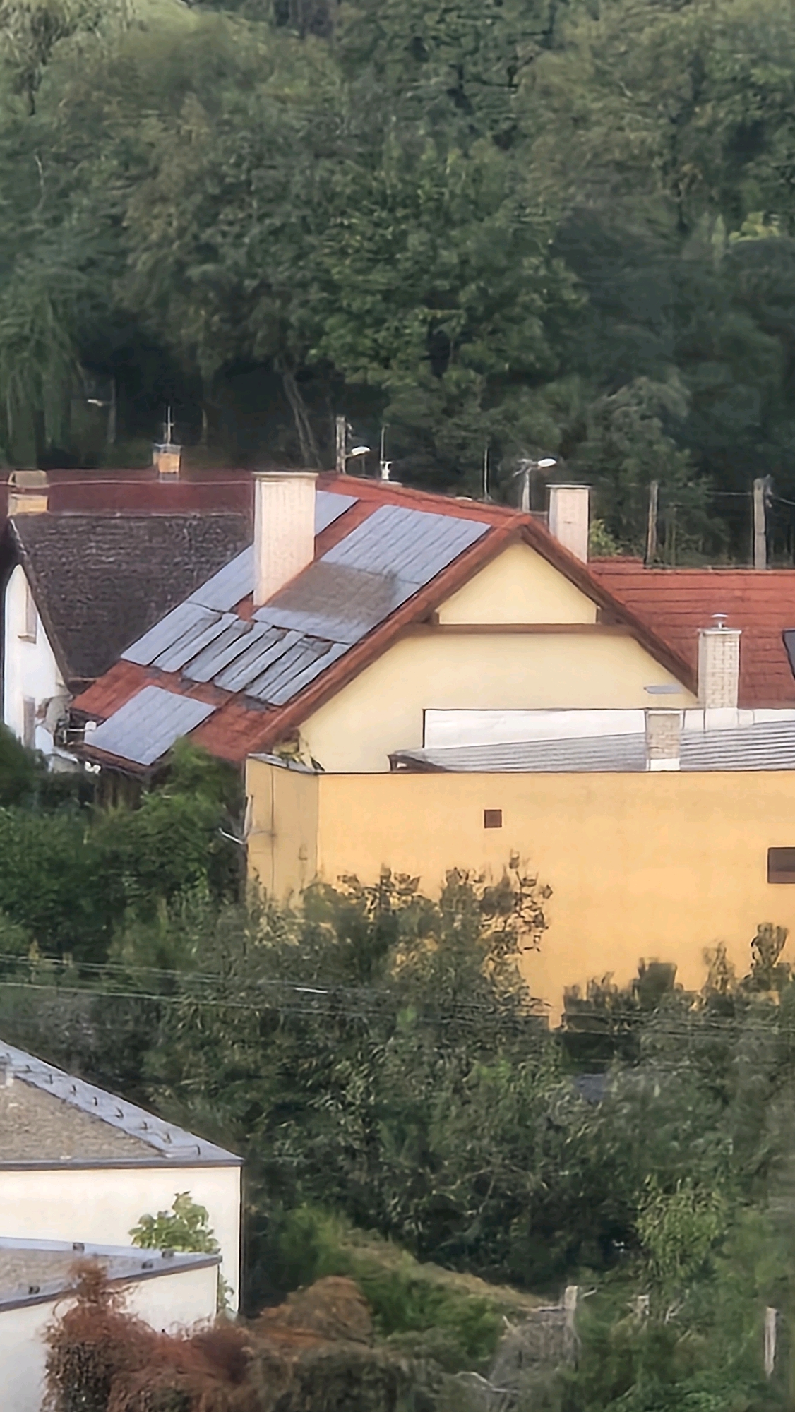 The image shows a residential area with houses and trees. The houses have red tile roofs, and some have solar panels installed. There are chimneys on some of the roofs. The houses are surrounded by lush green trees and vegetation. The overall scene suggests a suburban or rural setting within the city of Prešov, Slovakia. The lighting indicates it might be daytime, possibly late afternoon due to the warm tone of the houses. No people or specific actions are visible in the image.