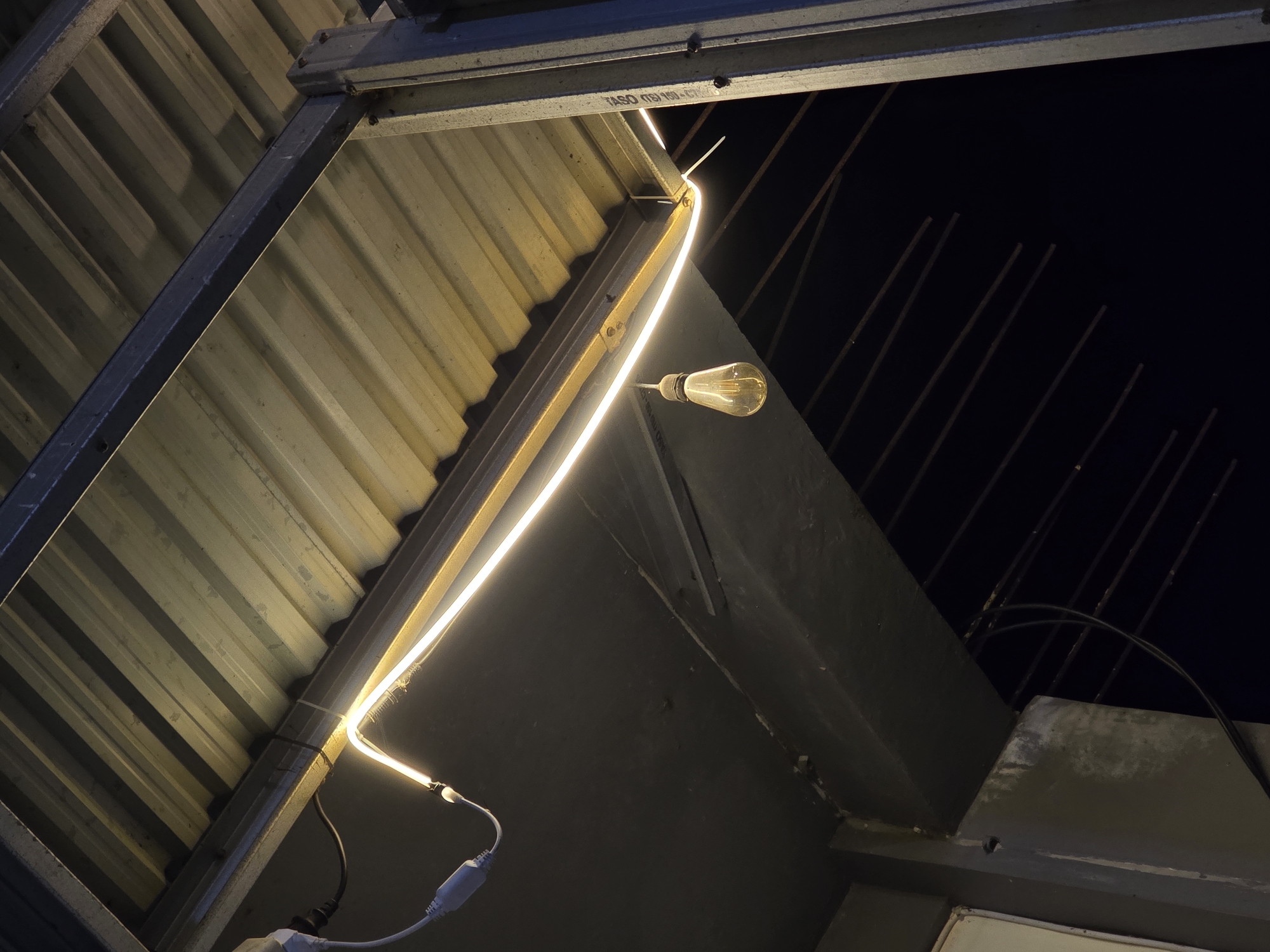 The image captures an outdoor scene at night, likely under a covered structure. A corrugated metal roof is visible, supported by metal beams. A bright, white LED strip light is affixed to the underside of the roof, tracing a path along a horizontal beam. A single Edison-style incandescent light bulb hangs from a wire, suspended below the LED strip and the roof. The background is dark, suggesting the night sky, with faint horizontal lines that could be shadows or distant objects. There are no people or animals present in the image. The environment appears to be utilitarian, possibly a porch, walkway, or open-air shelter. The specific location in Dayah Kruet, Indonesia, is not visually discernible from the image itself; it is inferred information provided with the request.
