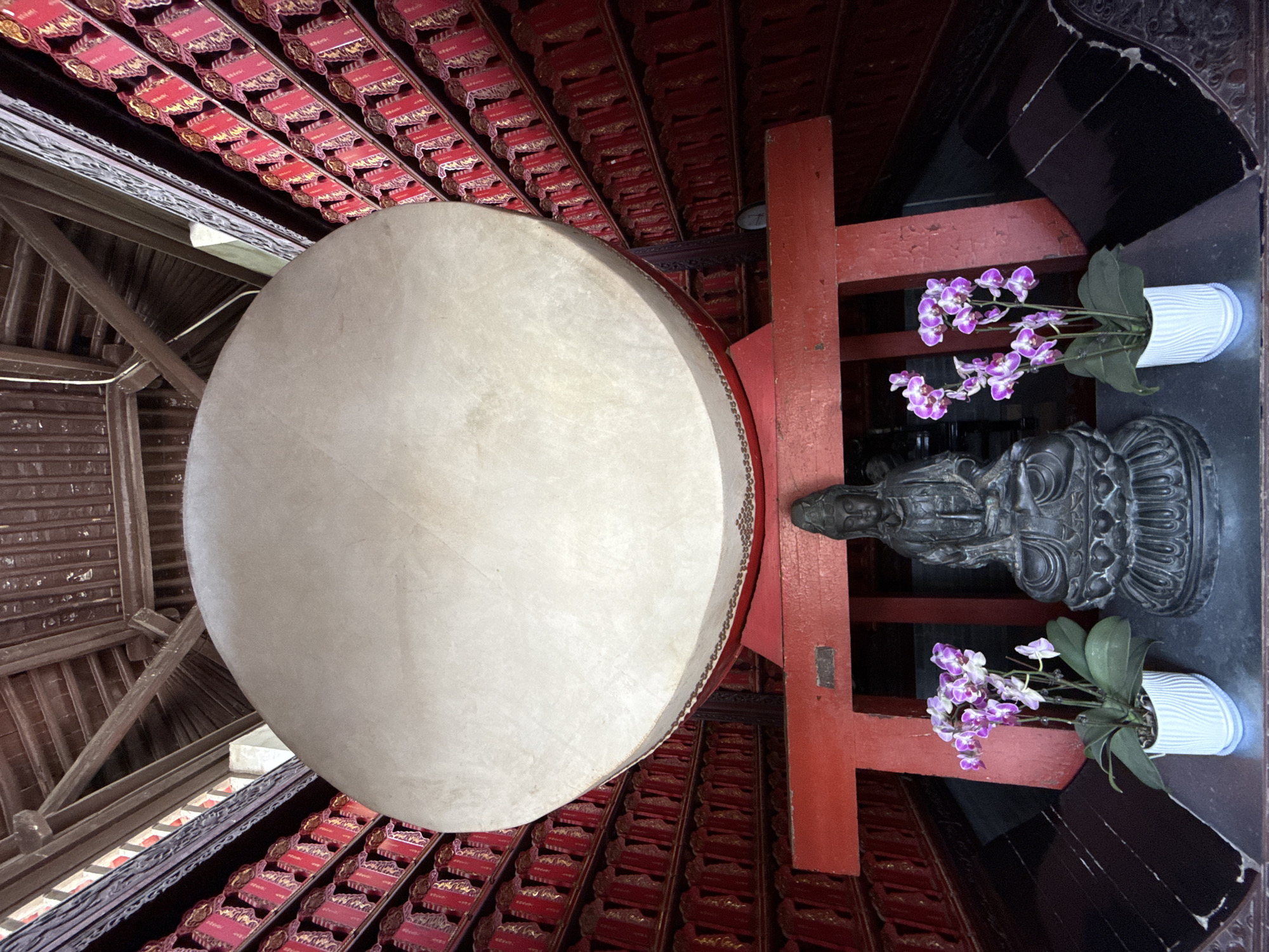 The image captures an interior scene in Yanta District, China. Dominating the view is a large, round drum suspended from the ceiling, the drum's surface being a pale, neutral color. The ceiling above features a patterned red and gold design, suggesting a traditional or religious architectural style. To the right of the drum, a red wooden structure stands near a dark stone statue, flanked by two small white pots containing purple orchids. The lighting is subdued, adding to the solemn atmosphere of the space.