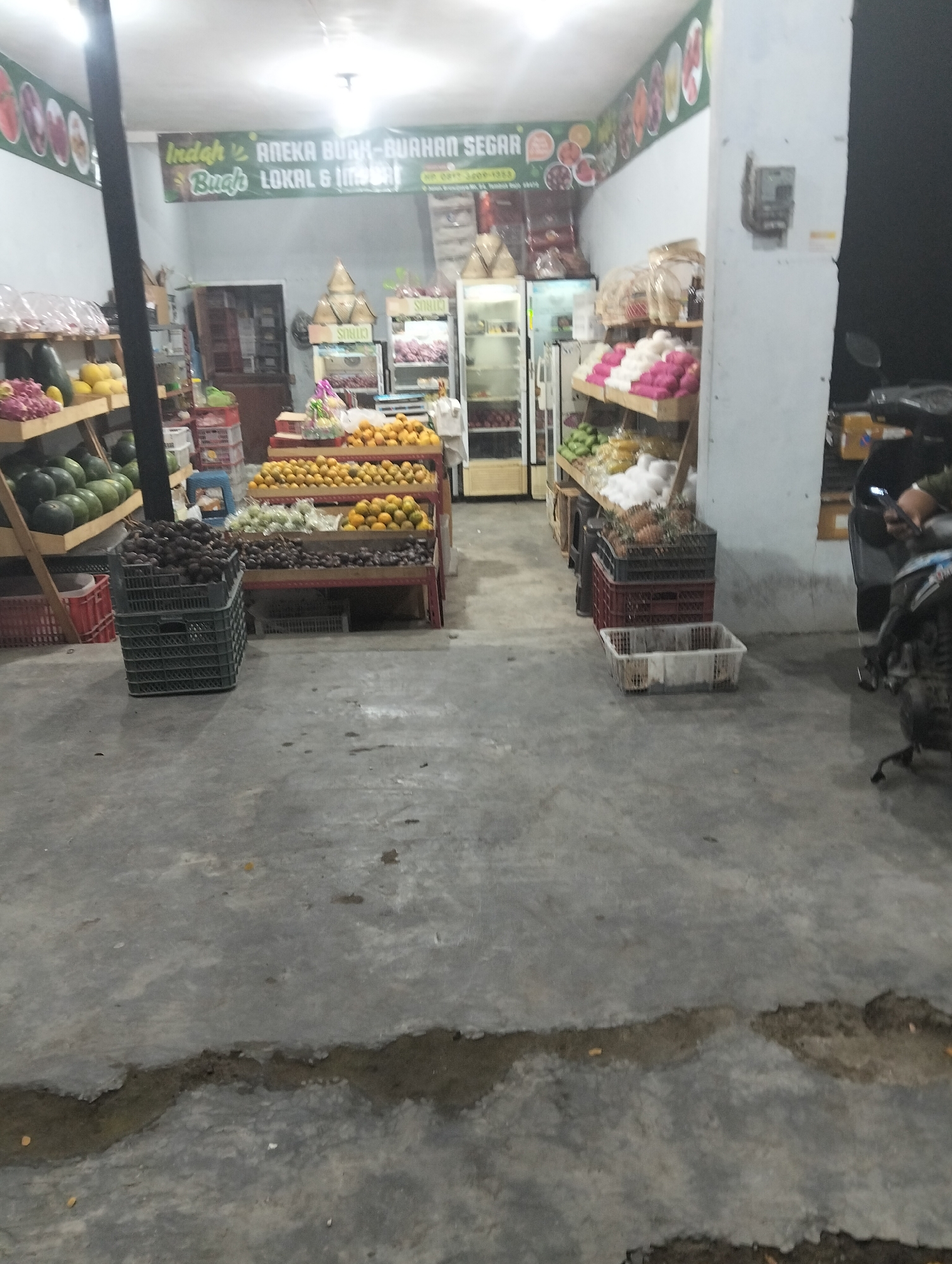 The image depicts the interior of a fruit and vegetable store in Muncar, Indonesia. The store is stocked with various fruits and vegetables displayed on shelves and in crates. In the foreground, a concrete floor is visible, with some cracks and debris. To the left, a shelf holds a variety of fruits, including what appear to be watermelons and smaller green fruits. Further back, more shelves display oranges and other produce. Behind the displays, a door leads to another room. On the right side of the image, a motorcycle is parked, with a person partially visible, possibly a customer or employee. The store appears to be dimly lit, with artificial lighting from above. A banner with Indonesian text, likely advertising the store's offerings, is hung on the wall above the produce. The overall impression is of a local market stall.