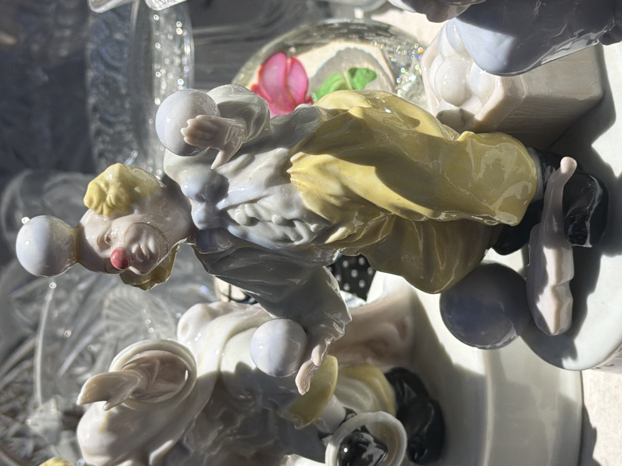 The image displays a close-up view of ceramic figurines of clowns, likely part of a collection or display. In the foreground, a clown figurine is depicted mid-performance, balancing a white ball on its head and holding two other white balls in its hands. The clown wears a yellow outfit with a white shirt and red nose. Behind this central figure, another clown figurine is partially visible, reaching out its hand. The figurines are set against a backdrop of what appears to be crystal decorative items, including a glass bowl with a pink and green floral design. The lighting suggests an indoor setting, possibly a shelf or display case, with sunlight casting shadows. The image provides no information about its location in Tel-Aviv, Israel.