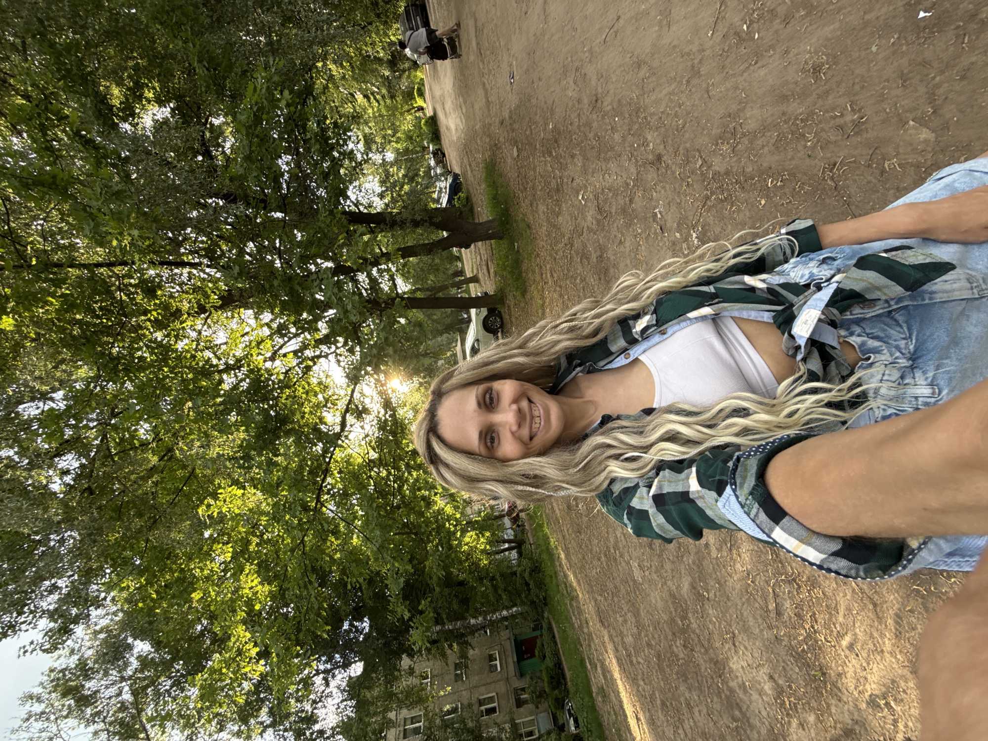 A medium shot shows a woman in Rusanovo, Russia. She is smiling at the camera. She wears a white tank top under a blue denim jacket and green plaid shirt. The ground is dirt and gravel. Behind her, a large tree and part of a building are visible. In the background, multiple parked cars are seen. The scene is lit by sunlight filtering through the trees.