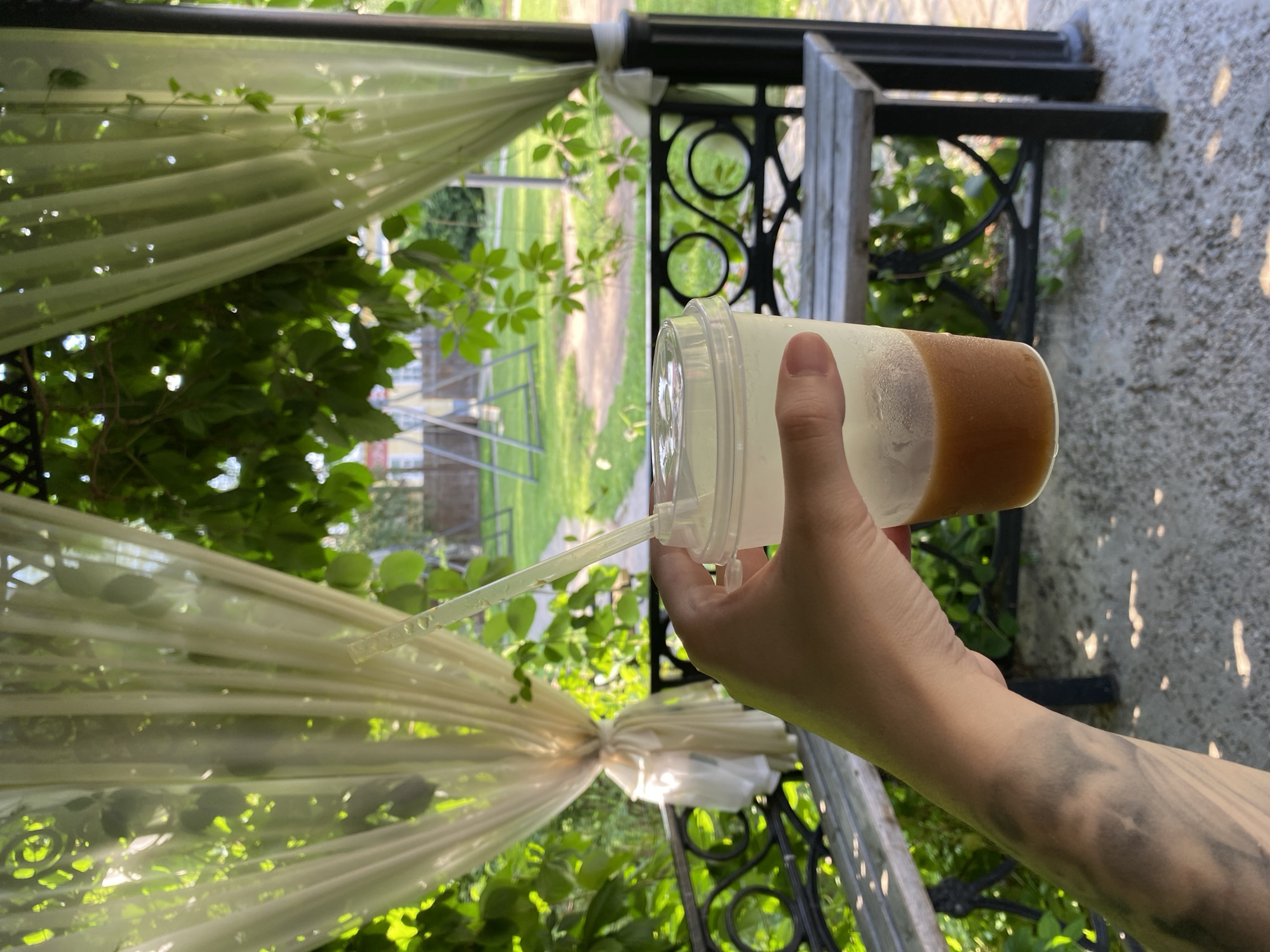 A hand holds a clear plastic cup with a light brown liquid and a straw. The cup is partially filled, and condensation is visible on its exterior. The background shows a gazebo with sheer curtains, lush green foliage, and outdoor furniture, including a wooden bench with decorative metal railings. Beyond the gazebo, a grassy area with a dirt path and playground equipment can be seen. The image appears to be taken outdoors on a sunny day. The location is identified as Votkinsk, Russia.