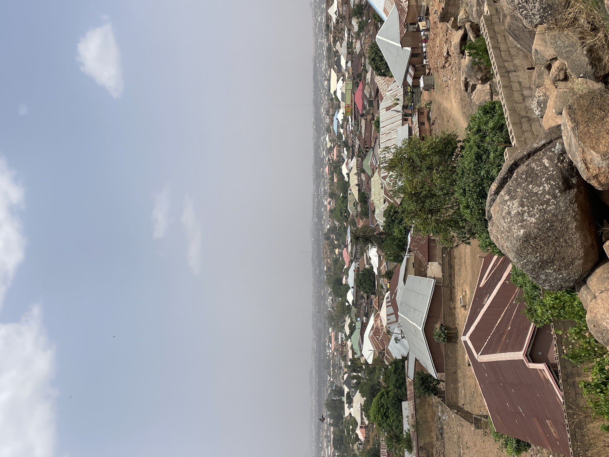 The image shows an elevated view of a section of Jos, Nigeria, with a hazy sky above. In the foreground, there are large rock formations. Houses with various roofing materials are clustered together, extending into the distance. A block wall can be seen leading into the buildings. The overall environment appears dense and residential.