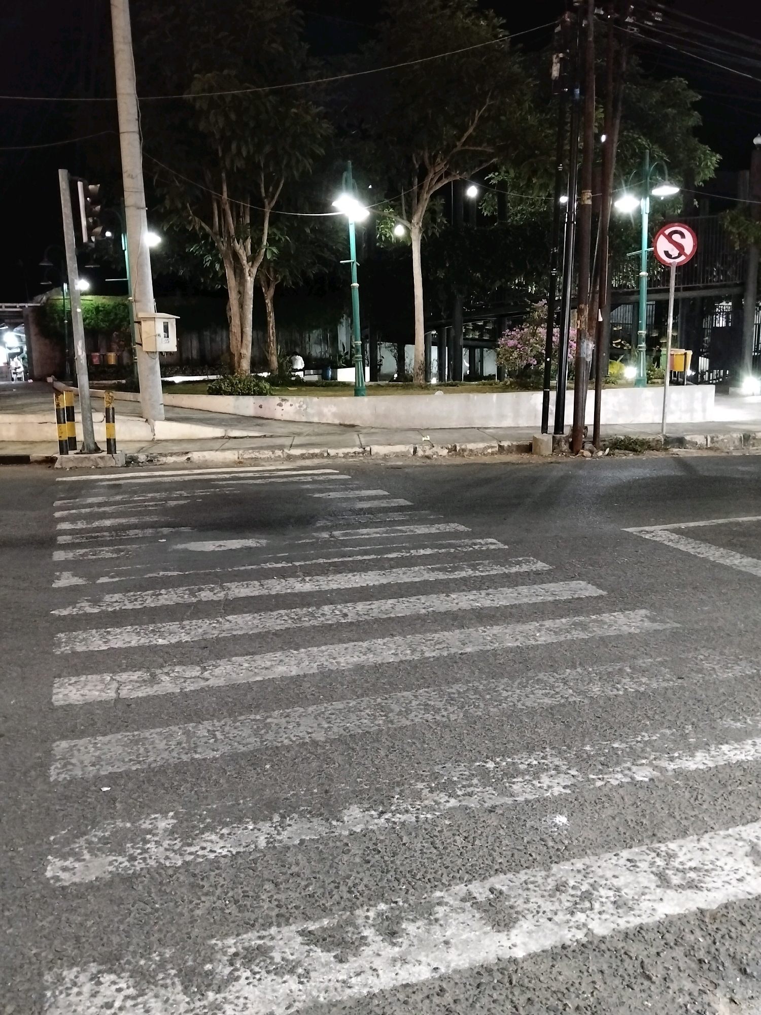 The image shows a crosswalk at night in the city of Purworejo, Indonesia. The crosswalk markings are white stripes on a dark asphalt road. In the background, there are trees, streetlights, and some buildings. A "no stopping" sign with the letter "S" inside a red circle is visible on the right side of the image. There are no people visible in the immediate vicinity of the crosswalk. The overall scene suggests a public space, possibly near a park or commercial area, that is lit by artificial lighting for nighttime visibility.
