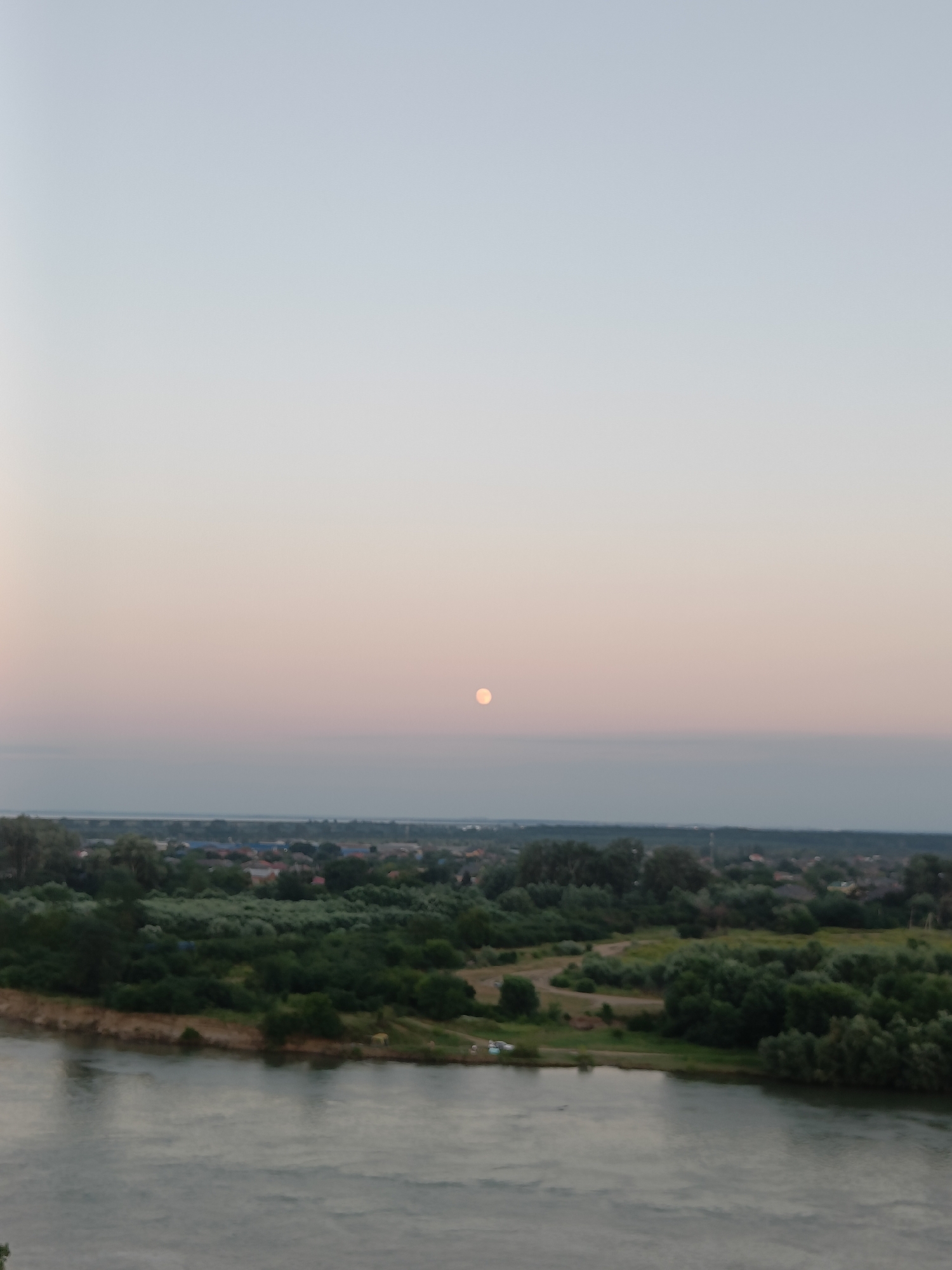 The image depicts a serene landscape under a twilight sky, possibly taken in the city of Krasnodar, Russia. The sky exhibits a soft gradient of pale blue and pink hues, with a full moon visible as a luminous orb near the horizon. Below the sky, a distant horizon line is marked by a hazy band, suggesting a body of water or a low-lying geographical feature.
The midground of the scene is dominated by a lush expanse of green trees and foliage, interspersed with what appear to be scattered buildings with red roofs, indicating a residential area. The vegetation is dense and varied, with trees of different sizes and shapes forming a rich tapestry of green. A notable feature is a wide river in the foreground, its surface reflecting the subdued light of the sky, creating a calm and slightly rippled texture. The riverbank is lined with greenery and a sandy or earthy embankment.
In the distance, beyond the immediate trees and settlement, the landscape stretches out, with hints of more dispersed buildings and vegetation. There are no discernible individuals or specific actions taking place within the frame, contributing to the overall sense of stillness and natural beauty. The image captures a moment of transition between day and night, highlighting the tranquil beauty of the natural and semi-urban environment.