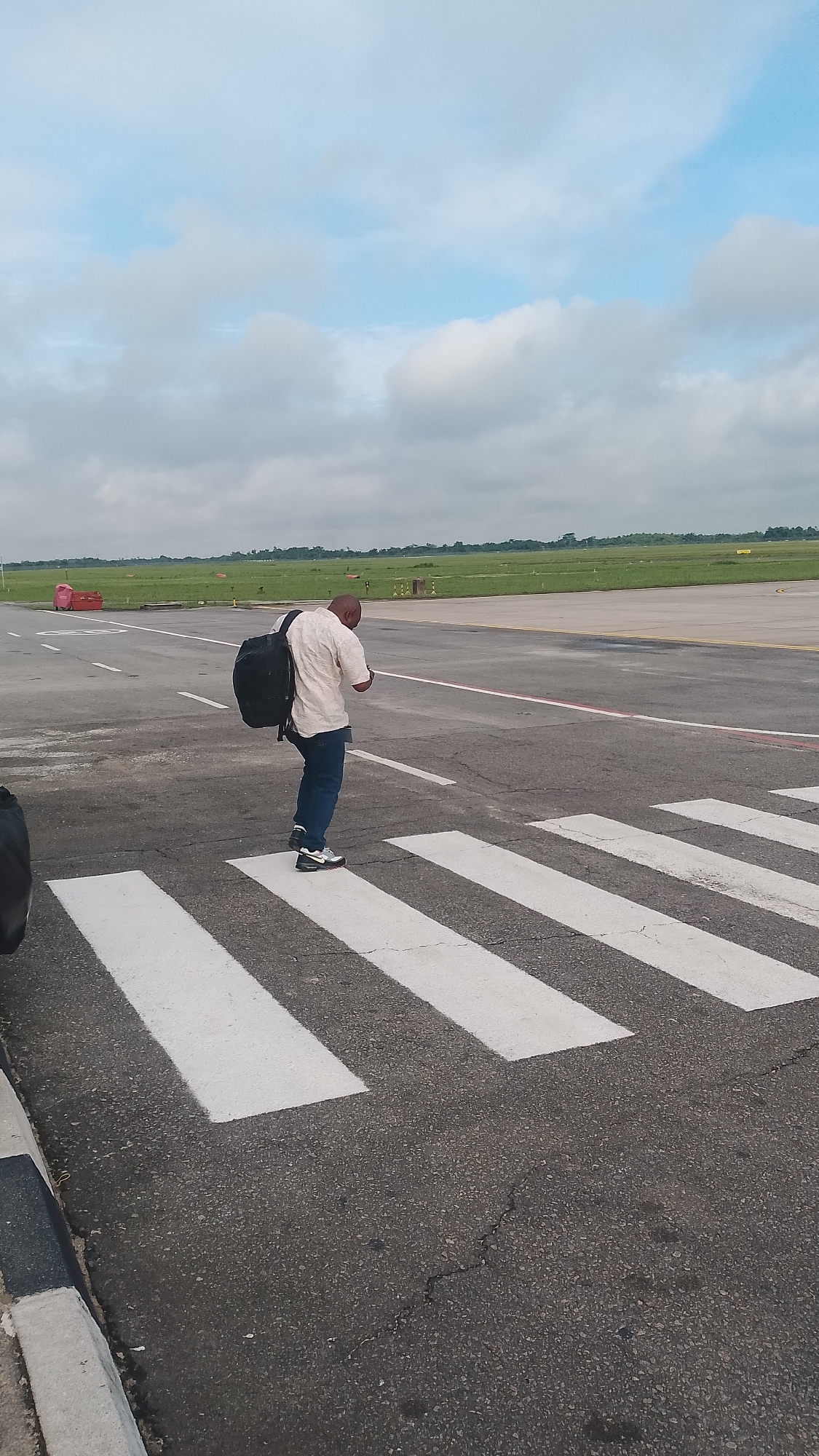 A person with a backpack is walking across a pedestrian crossing at what appears to be an airport in Orerokpe, Nigeria. The person is wearing a light-colored shirt and blue jeans. The sky is cloudy with patches of blue sky visible. The ground is asphalt with white pedestrian crossing stripes. In the background, there is a green grassy field and what looks like an airport runway. There is also a red object, possibly equipment, in the far background. The scene appears to be an outdoor area on a cloudy day.