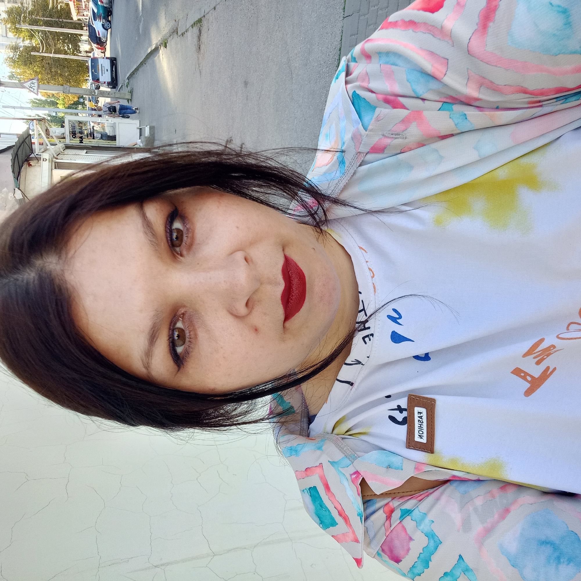 The image is a close-up selfie of a woman in Kropyvnytskyi, Ukraine, taken outdoors during the day. The woman has dark hair, brown eyes, and is wearing dark red lipstick. She is wearing a white t-shirt with colorful graphics and a patterned, open jacket over it. The background shows a street scene with a sidewalk, buildings, parked cars, and trees. The woman is standing on a sidewalk next to a building with a cracked white wall. The street is paved, and there are power lines overhead. A woman can be seen walking away from the camera on the sidewalk in the mid-ground. The overall lighting suggests it is daytime.