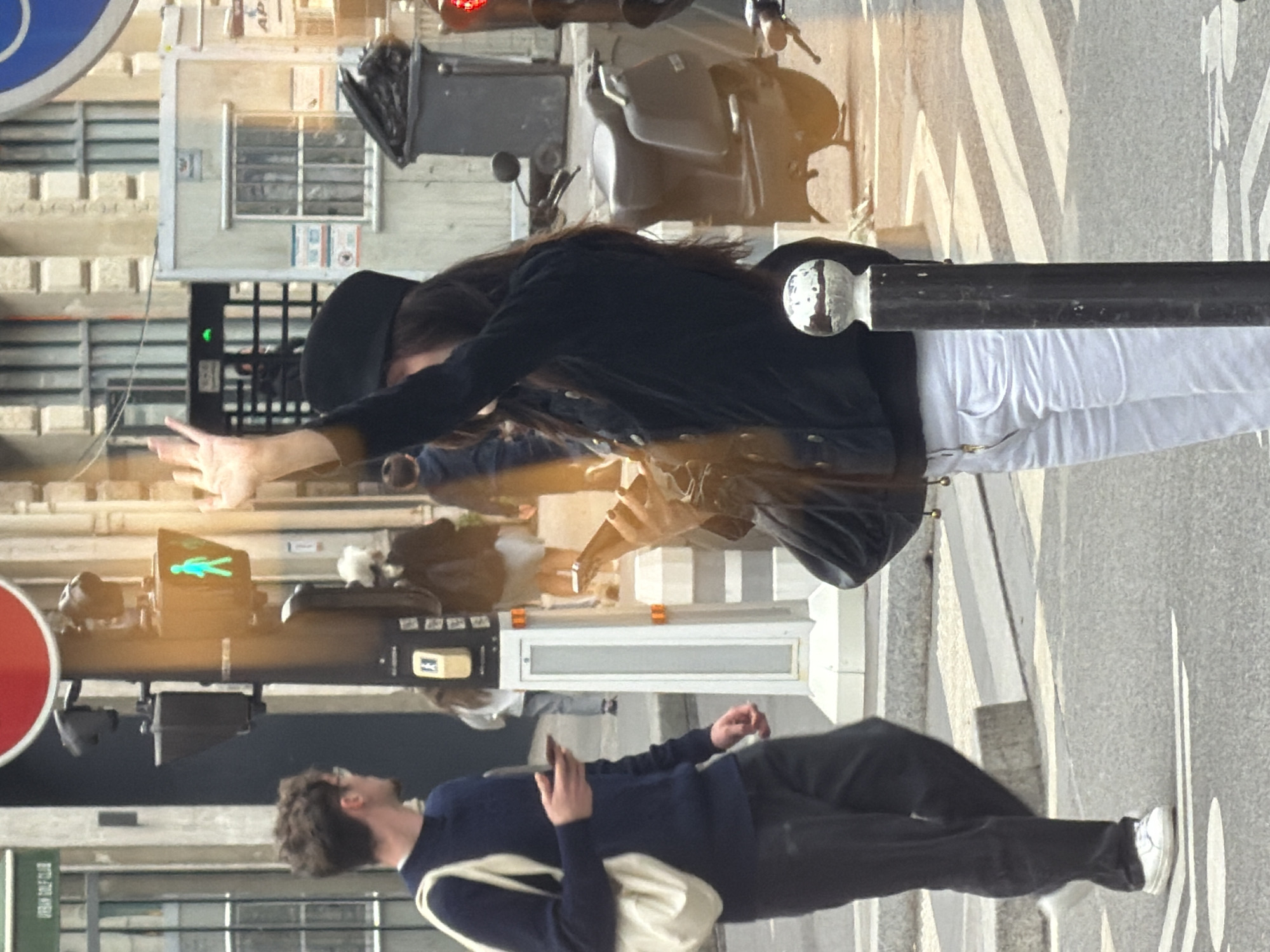 The image shows a street scene in Paris, France. A pedestrian crosswalk is visible. A person in a black jacket, white pants, and a black hat is crossing the street, their arms extended. A man with dark hair wearing a blue sweater and black pants stands at the crosswalk, looking in the direction of the person crossing. A white bag is slung over his shoulder. A traffic light indicates it is safe to walk. A scooter is parked nearby. Buildings are visible in the background.
