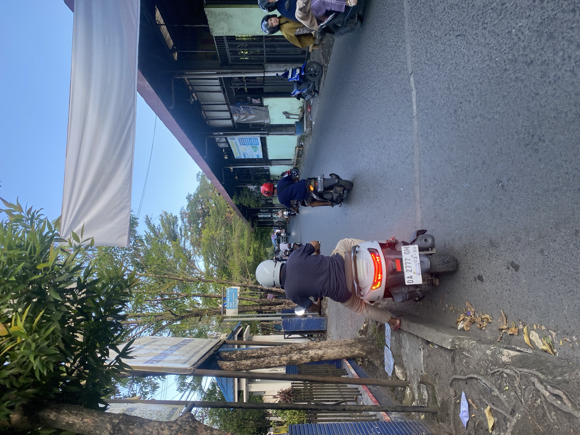In Desa Tunggul Irang, Indonesia, a street scene unfolds under a clear sky with a few trees lining the left side of the road. A man wearing a blue shirt and a white helmet is riding a white scooter, identified by the license plate DA 2777 QM. He is traveling down the asphalt road, which appears to be a public street. To the right of the road, a long, low building with a red-brown roof runs alongside the street. Several other motorcycles are visible in the background, some carrying passengers. The road surface shows signs of wear and tear, with some cracks visible. On the left side of the road, there is a sidewalk area with exposed tree roots and scattered leaves. A blue sign with white text is mounted on a pole near the trees. The overall impression is that of a typical day in a residential or commercial area of the town.