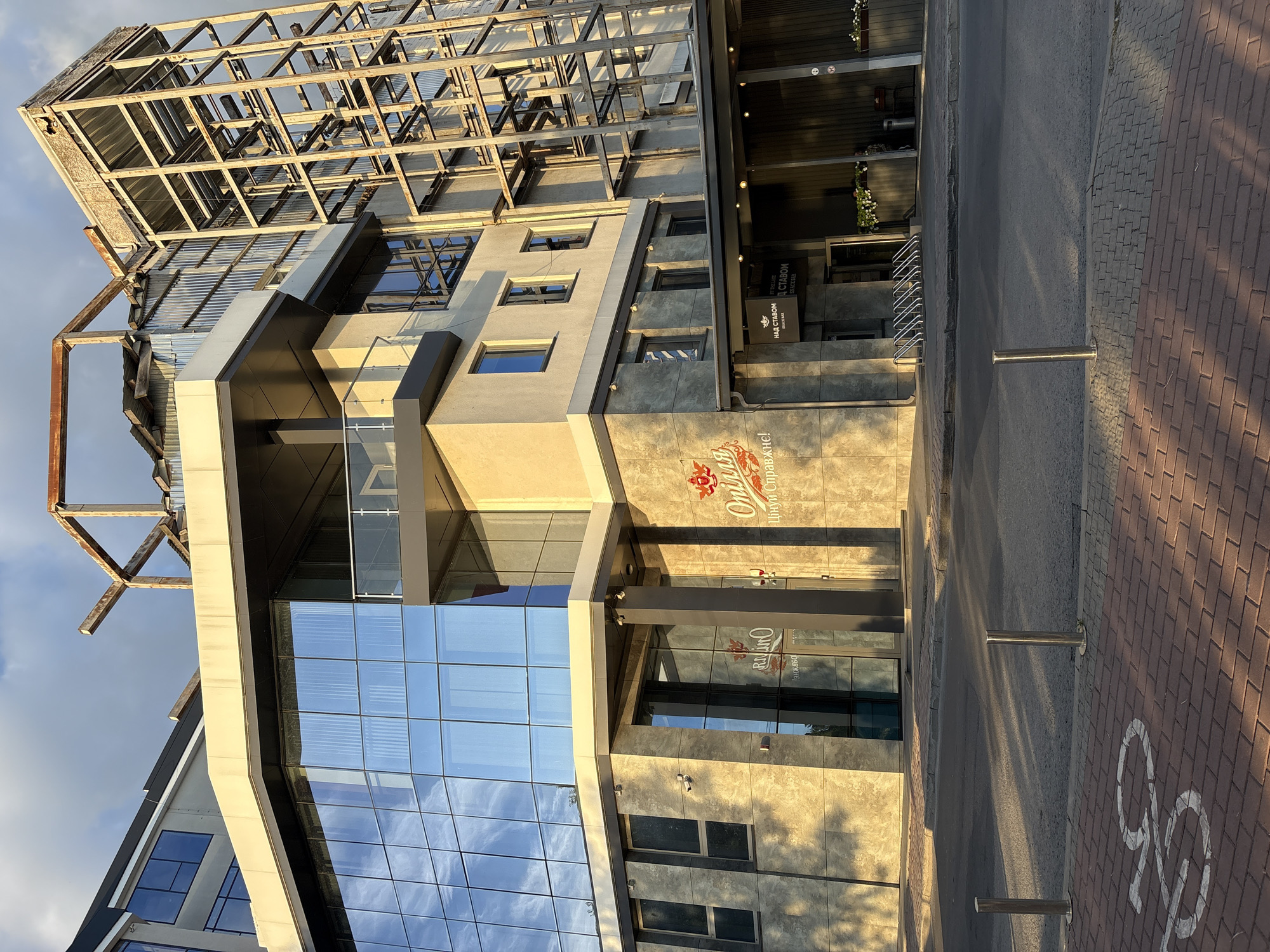 This is a full exterior shot of a modern building with a large glass facade in Ternopil, Ukraine. The building appears to be a business or restaurant establishment, as indicated by the signage on the exterior. The sign above the entrance reads "НАД СТАВОМ" which translates to "Over the Pond". Below that, it says "SNACK BAR SINCE 2018". Another sign on the glass facade reads "Опція" and "Ципу Справжнє!". The building features a mix of glass, concrete, and metal elements. To the right of the main building structure, a partially constructed or under-renovation metal frame is visible, suggesting ongoing development or damage. The sunlight casts long shadows across the pavement and the building itself, indicating it is either early morning or late afternoon. There are bollards and a bike rack in front of the building. The street in the foreground has a bicycle symbol painted on the brick pavement. No people are visible in the image.