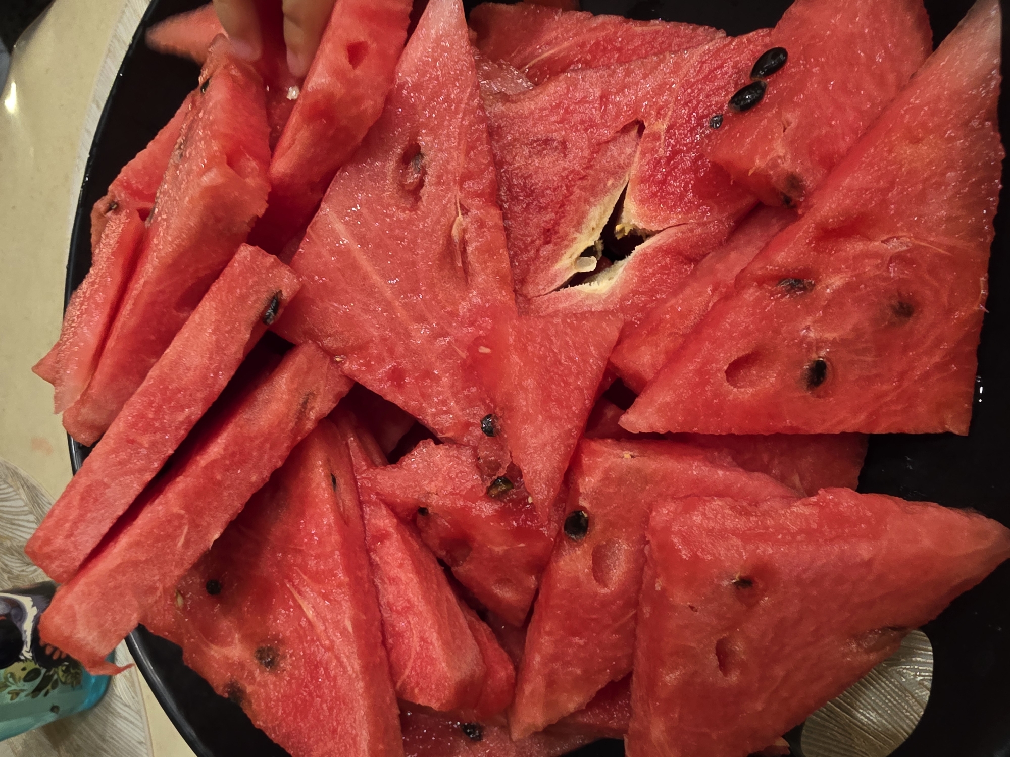 A close-up shot shows a pile of sliced watermelons in a black bowl. The watermelons are cut into triangular wedges and rectangular strips, with some pieces showing visible black seeds. The flesh of the watermelon is a vibrant red color, and some pieces appear moist, suggesting they are fresh. In the background, a glass with a pattern of birds and leaves is partially visible, along with a white textured surface, possibly a tablecloth. A hand is seen reaching into the bowl on the right side of the image, possibly taking a piece of watermelon. There is no discernible location in Saint Petersburg, Russia, as the image focuses solely on the food and its immediate surroundings.