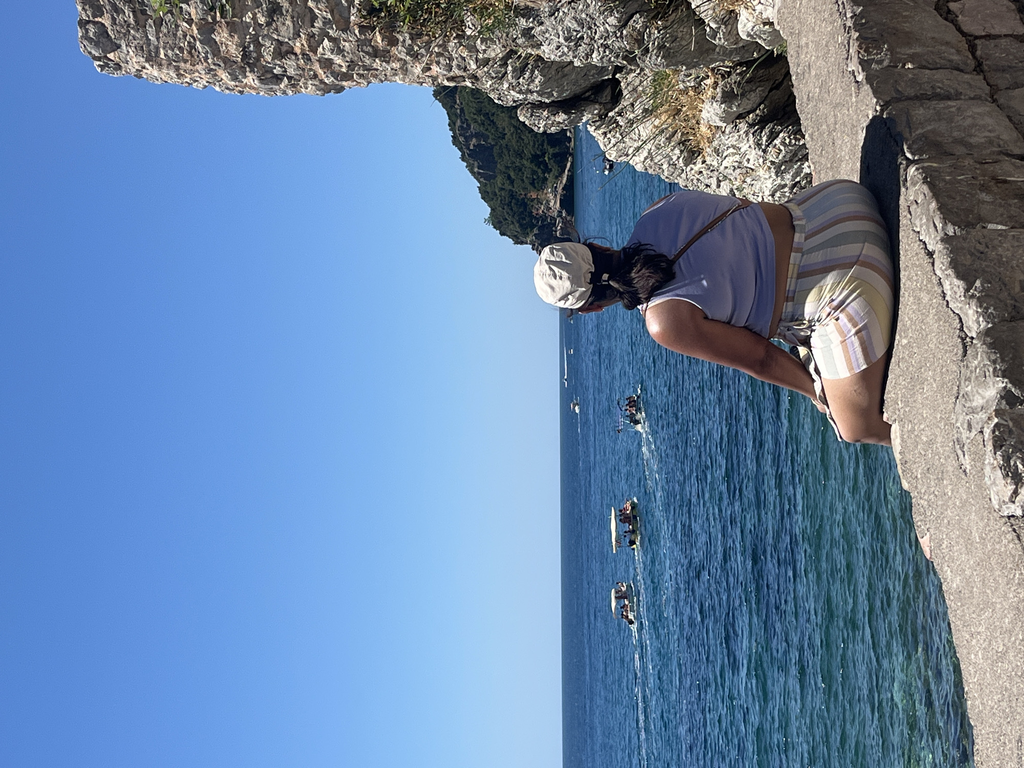 A woman wearing a light-colored top, striped shorts, and a hat stands on a stone ledge overlooking the Adriatic Sea in Budva, Montenegro. Several small boats traverse the blue water. The sky is clear blue. A rocky cliff overhangs the ledge on which the woman stands.
