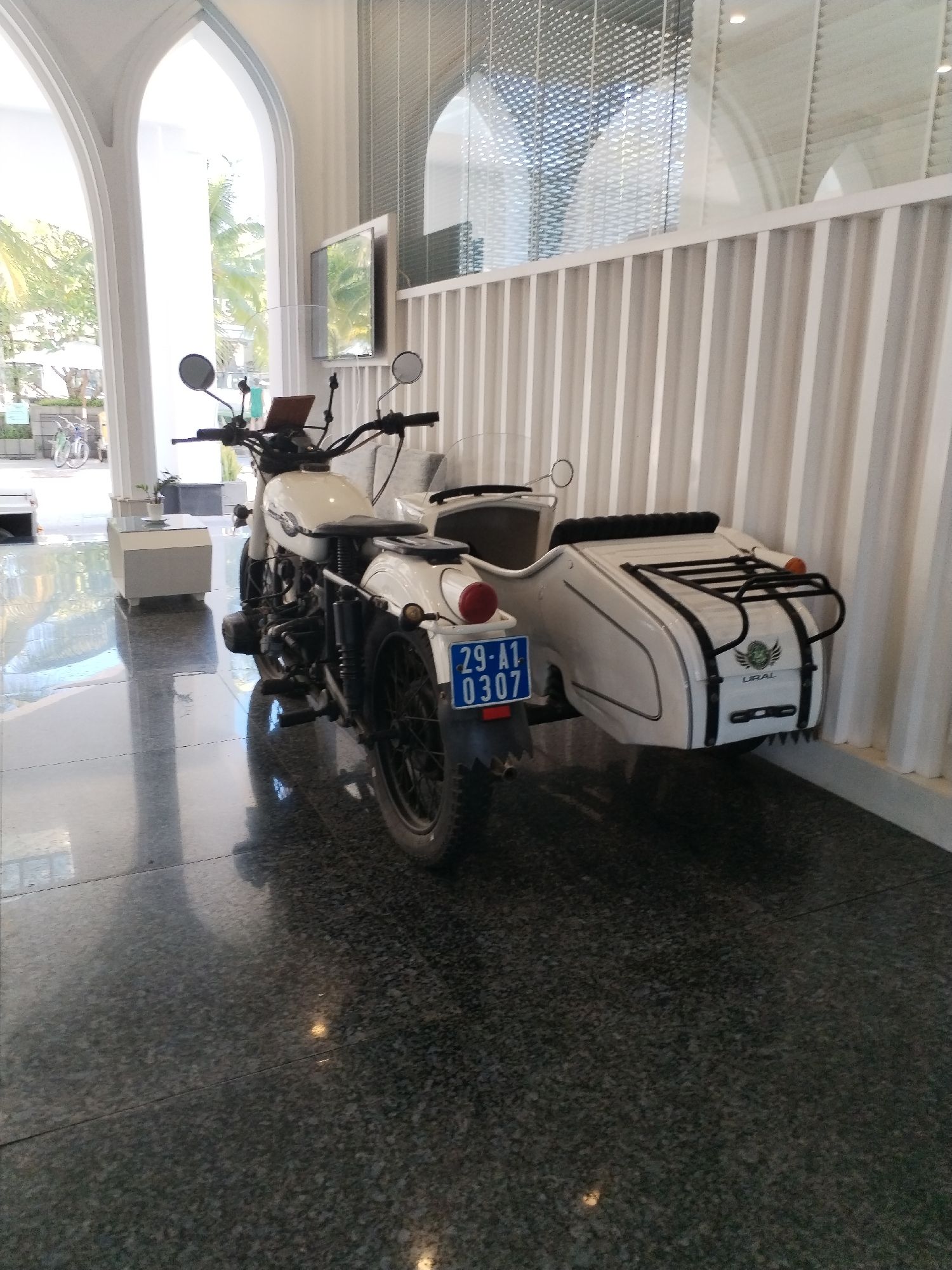 A white Ural motorcycle with a sidecar is parked inside a building with arched windows. The motorcycle has a blue license plate with white lettering that reads "29-A1 0307". The sidecar has a black luggage rack and the word "URAL" printed on its side. The floor is polished black granite. In the background, there is a white slatted wall and a television screen. Outside the arched windows, palm trees and bicycles are visible. The location is identified as Bac Nha Trang Ward, Vietnam. There are no people in the image.