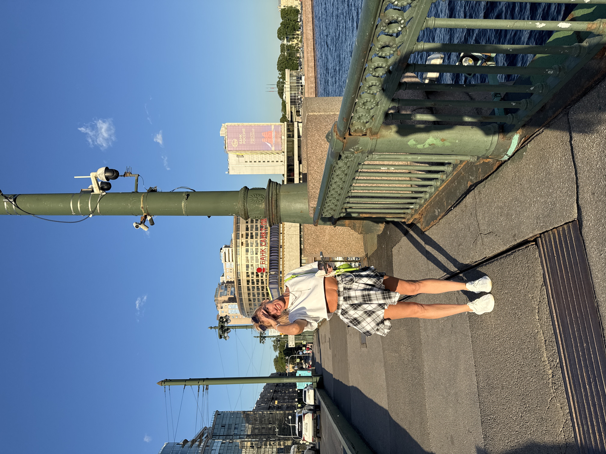 A woman with blonde hair stands on a bridge in Saint Petersburg, Russia. She is wearing a white crop top, a black and white plaid skirt, and white sneakers. A bright green bag is slung over her shoulder. She is shielding her eyes from the sun with her hand. In the background, there are buildings, cars, a body of water, and a large green lamppost with security cameras attached. The sky is a clear blue with a few small white clouds. The scene appears to be illuminated by bright sunlight, casting shadows on the bridge.