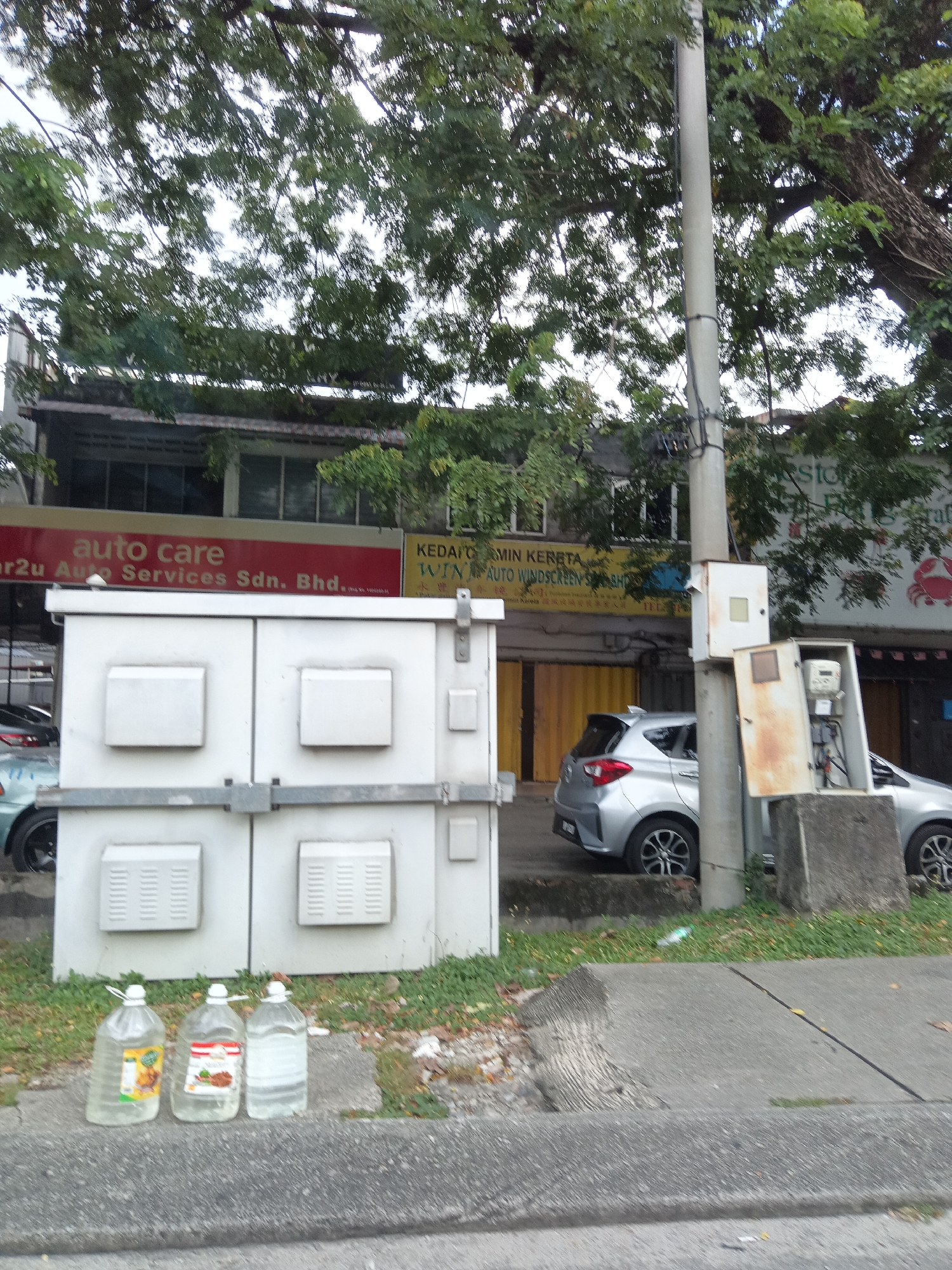 In Kuala Lumpur, Malaysia, on a street with trees overhead and a sidewalk, a white utility box with multiple compartments stands near a gray car. Three large plastic bottles, two with labels and one clear, are placed on the curb in front of the utility box. A gray pole with wires and two smaller utility boxes is positioned to the right of the main utility box. Buildings with signs are visible in the background.