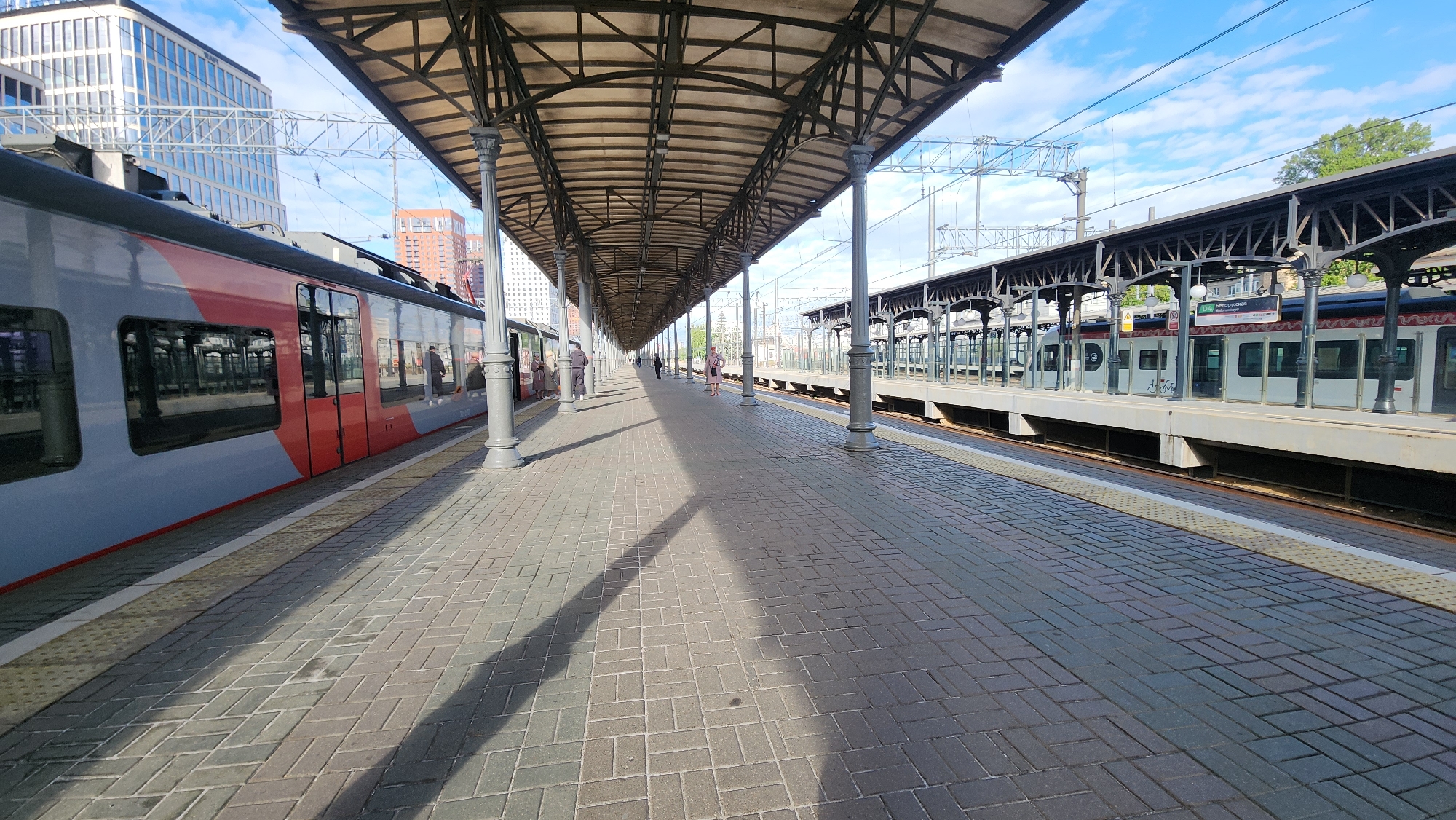 The image shows a train station platform in an urban area. The platform is paved with rectangular gray bricks and is covered by a metal and glass canopy supported by ornate gray pillars. On the left side of the platform is a train with a gray and red exterior. Passengers are visible inside the train through the windows. On the right side, another train is parked along a raised platform. A few people are seen walking on the platform, positioned towards the center of the frame. Overhead electrical lines are visible above the canopy structure. In the background, buildings of various heights and architectural styles can be seen under a partly cloudy sky. The location appears to be in the city of Unknown, Unknown.
