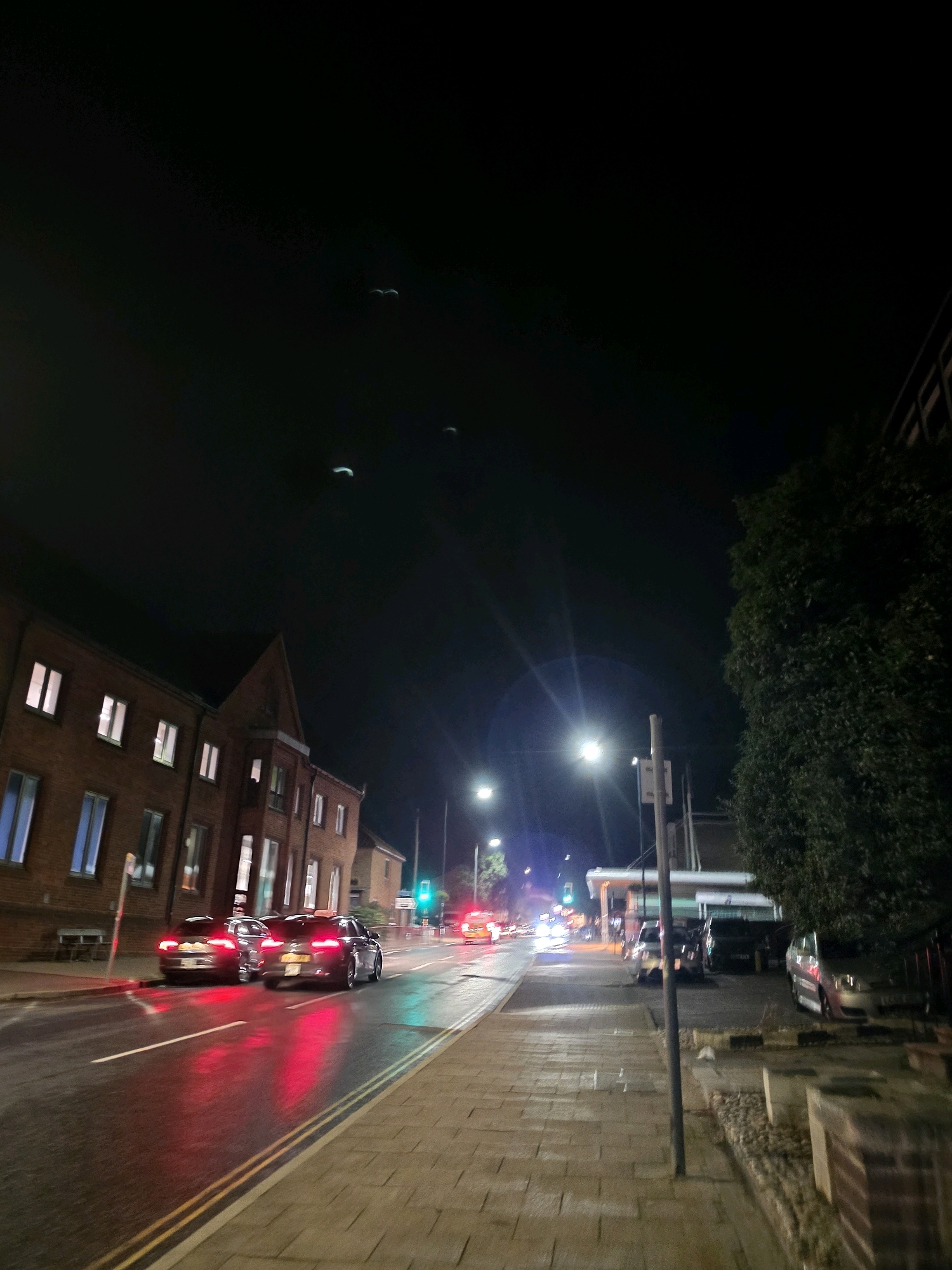 The image shows a street scene at night in West Suffolk, United Kingdom. The street is wet, reflecting the red and green lights from vehicles and traffic signals. There are several cars on the road, some with their brake lights illuminated. Buildings line the left side of the street, appearing to be residential or commercial properties with illuminated windows. A large tree with dense foliage is visible on the right side of the image, obscuring part of the sky and a building behind it. Streetlights are illuminating the scene. In the sky, there appear to be three faint, crescent-shaped objects. The pavement is made of brick or stone pavers.