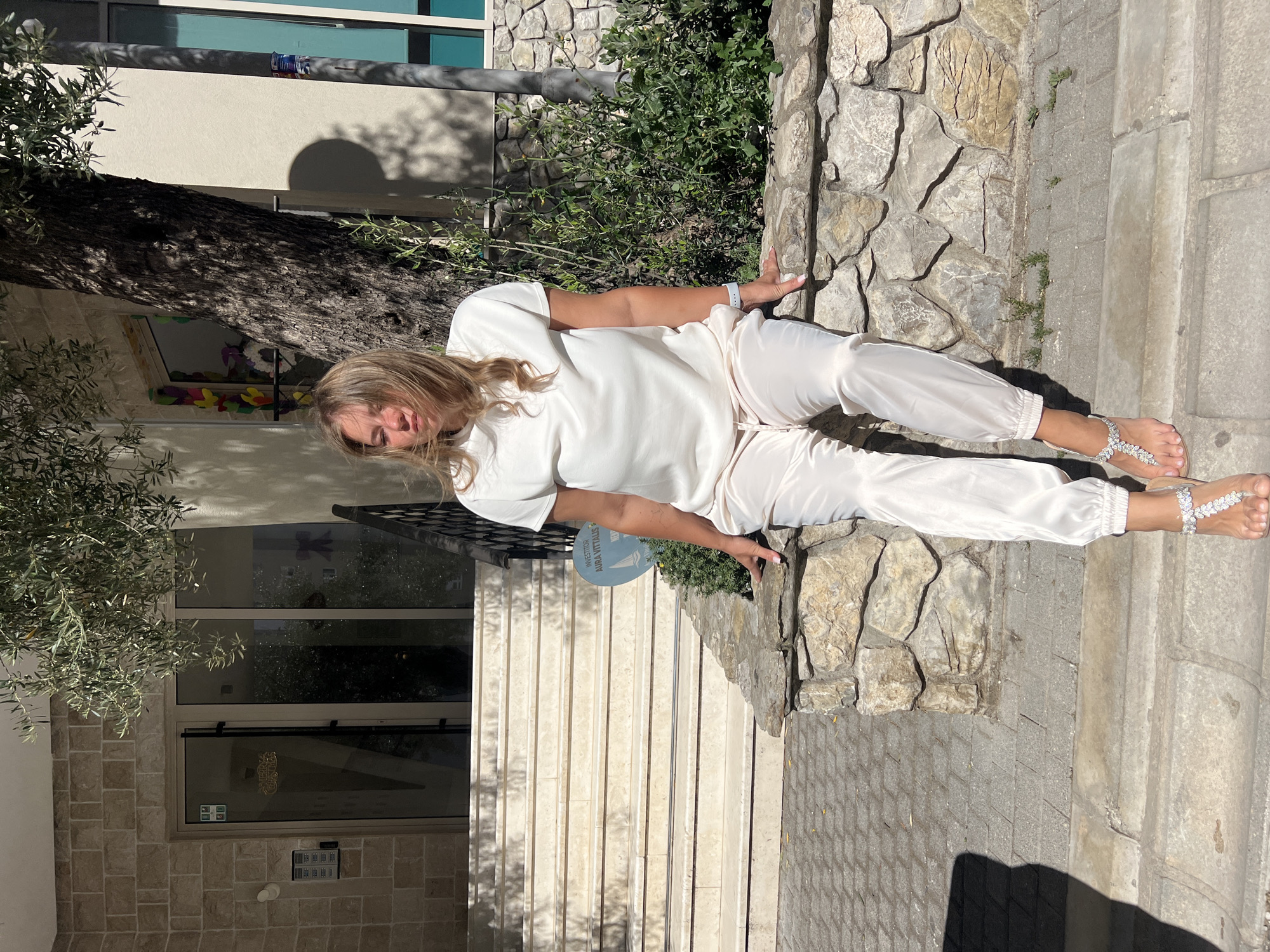 A woman is captured in a full-length, outdoor portrait, leaning casually against a textured stone wall. She has blonde hair and is wearing a matching light-colored (likely white or cream) short-sleeved top and loose-fitting pants, possibly satin or silk, with elasticized ankles. Her feet are adorned with decorative, possibly jeweled, flat sandals. A light-colored watch is visible on her left wrist. She is looking towards the camera with a relaxed expression.
The setting appears to be a sunny outdoor area, likely a pedestrian zone or courtyard adjacent to a building. Strong shadows cast by the woman and nearby trees indicate bright sunlight, suggesting midday or early afternoon. To the woman's left, a large tree with dark, gnarled bark (possibly an olive tree, given the provided location context of Budva, Montenegro) is prominent, with its leaves visible on the left side of the frame. Behind the woman, there's a stone wall made of irregular, light-colored blocks.
To the left of the tree and behind the woman, a light-colored building with windows is visible, featuring a tiled facade at the bottom. Below the building's entrance, a security panel is mounted on the wall. A set of light-colored steps leads up to what appears to be the entrance of the building. A blue, circular sign is affixed near the steps, displaying the text "INVESTITOR AURA VITALIS". The ground beneath her is paved with grey interlocking bricks, and a concrete curb or edge is visible to her right. Lush green foliage grows in various spots along the stone wall and in the background.