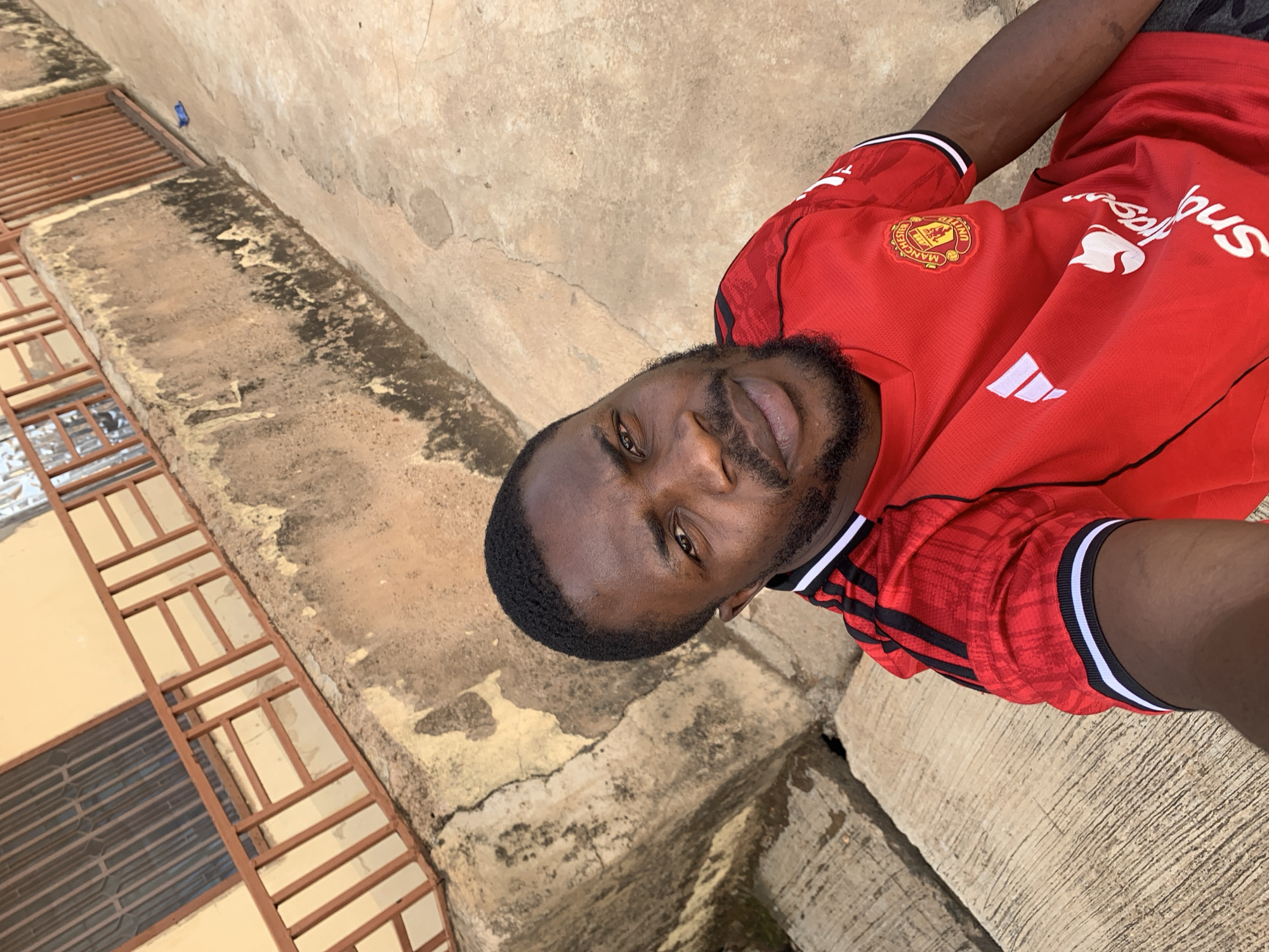 A man wearing a red Manchester United soccer jersey is pictured in Rafin Par, Nigeria. He stands near a building with a brown-and-tan concrete exterior. The building has metal window bars. The ground appears to be concrete as well, cracked and weathered.