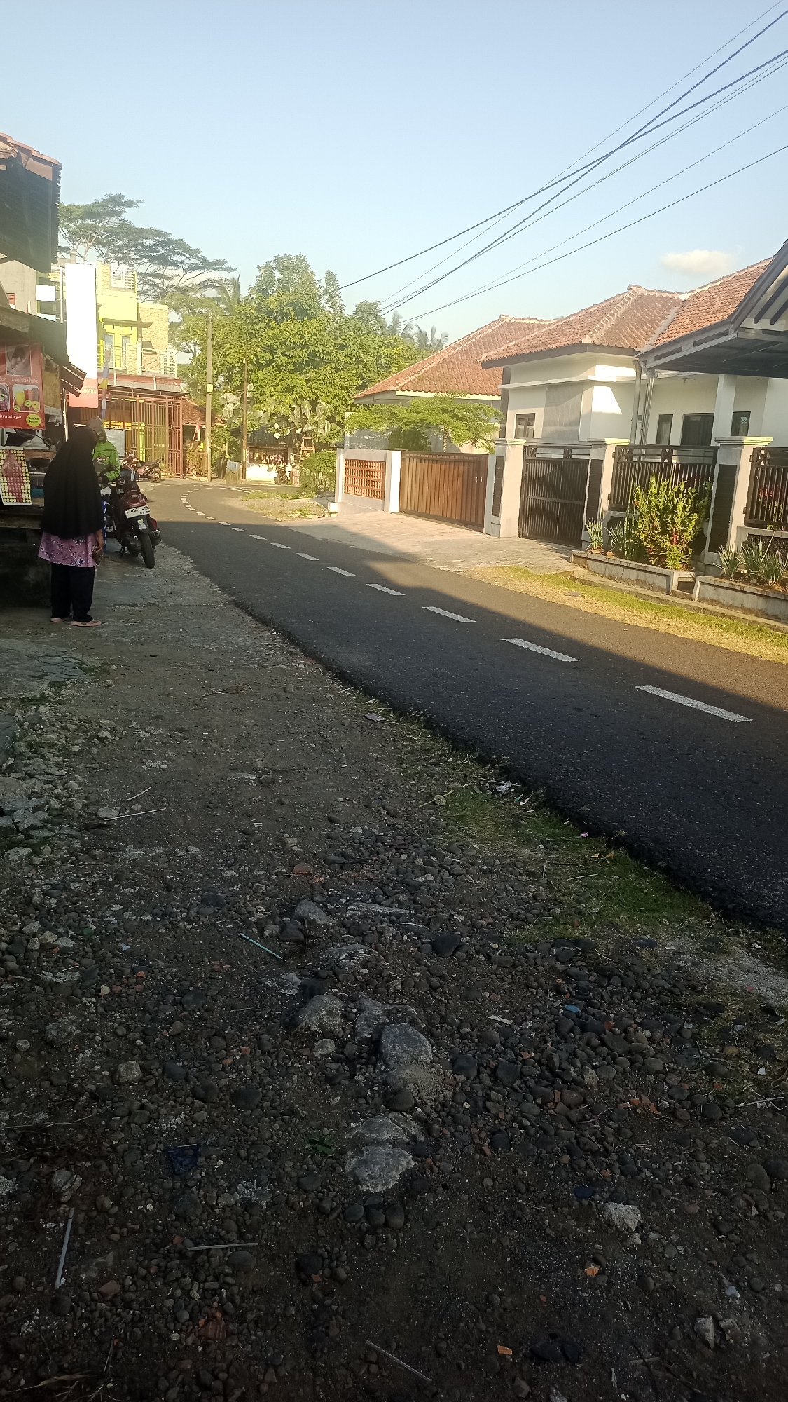 The image depicts a street scene in Pakemitan, Indonesia, likely during the early morning or late afternoon, given the low angle of the sunlight creating long shadows. The road, paved with asphalt, is marked with white dashed lines. To the left of the road, a person wearing a black hijab and a patterned pink and white shirt stands near a motorcycle. Adjacent to them is a small structure with a sign displaying colorful images, suggesting a small shop or food stall. The foreground of the image is dominated by a rough, gravelly terrain with scattered rocks and some debris.
On the right side of the road, several houses are visible. These houses feature terracotta tiled roofs and are separated by fences made of metal and wood. Some houses have well-maintained gardens with plants and decorative elements. The overall environment appears to be a residential area within a town or city, with a clear sky overhead. Power lines are strung across the upper portion of the image. The scene conveys a sense of everyday life in a tropical urban setting.