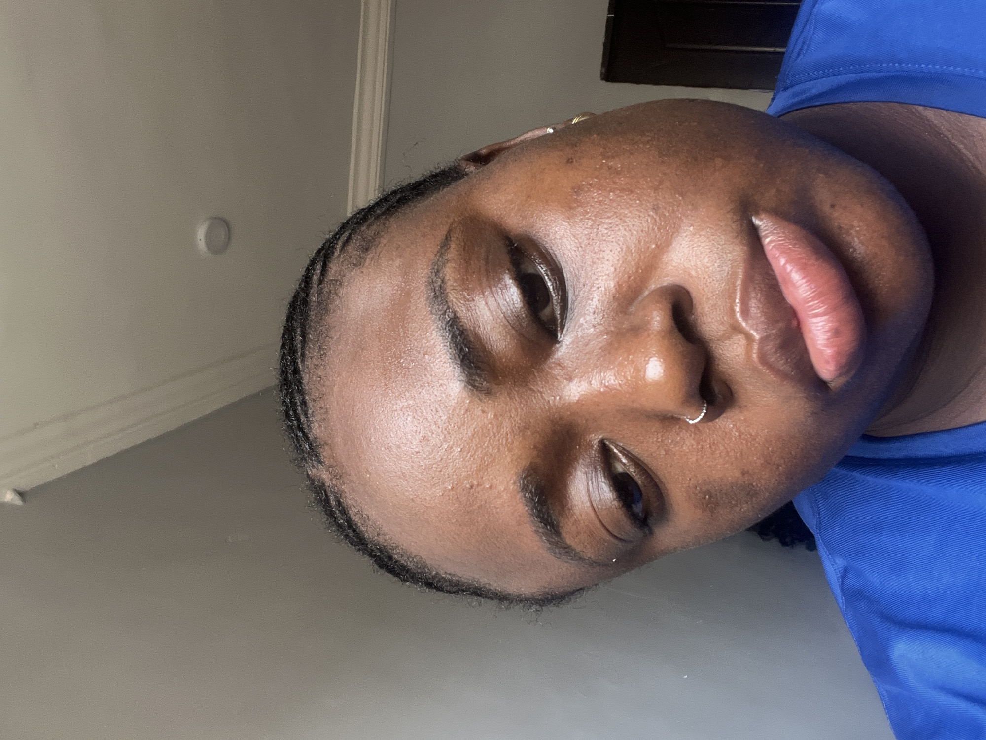 A close-up, angled portrait of a Black woman's face. She has dark skin, a nose ring, and small braids. Her lips appear glossy. She is wearing a blue top. The background consists of a light gray wall and a glimpse of a dark object near the top right corner, possibly a window or furniture. No evidence indicates the location is Kubwa, Nigeria.