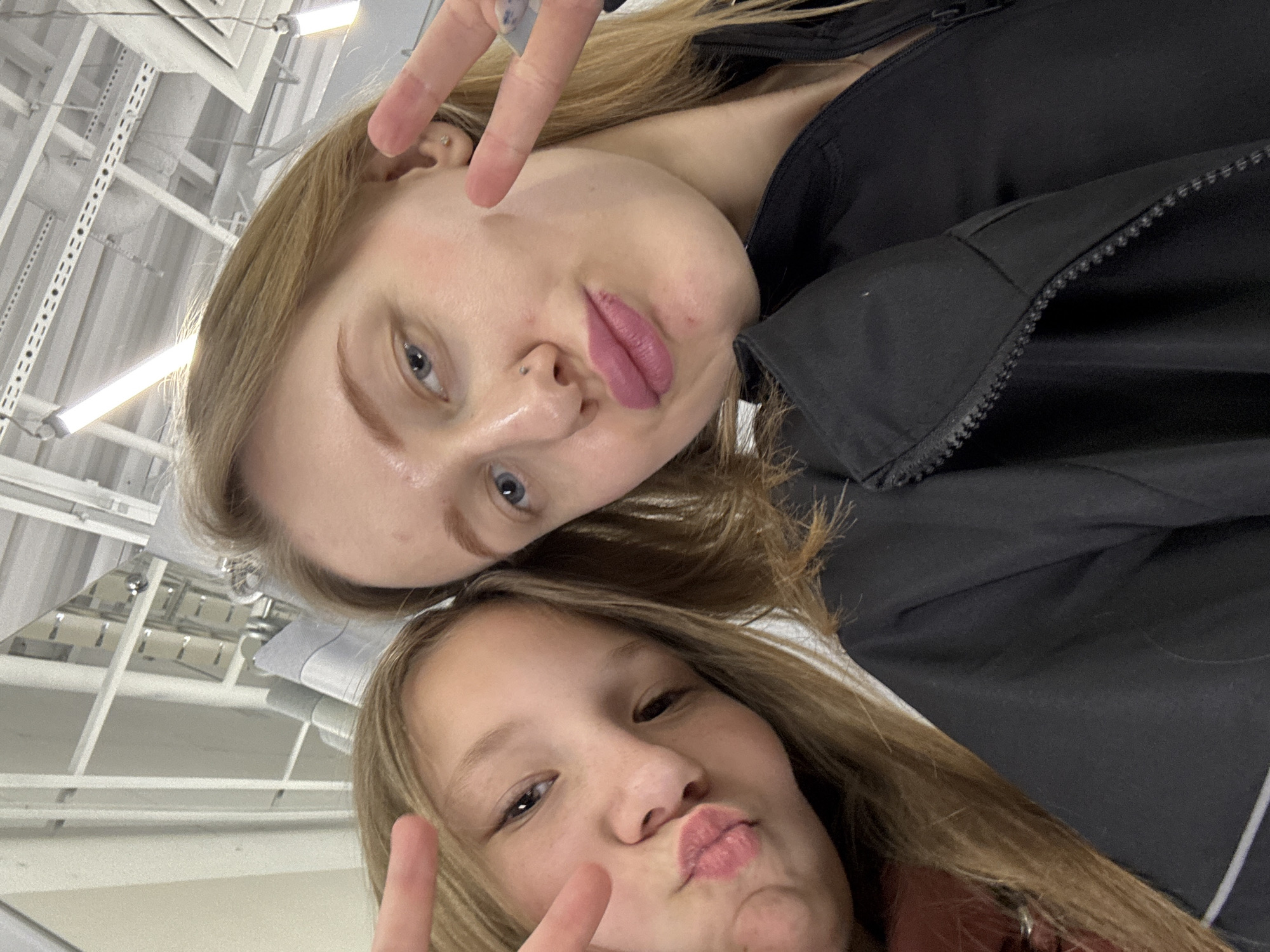 Two young women are posing for a selfie indoors. They both have fair skin, blonde hair, and are making kissing faces while holding up the peace sign with their fingers. The woman in the upper part of the frame is wearing a black jacket and has a nose piercing. The ceiling above them is visible, showing fluorescent lighting and support structures. The photograph has a slightly tilted perspective. There is no information to specify if the image was taken in Ovoshchnoy, Russia.