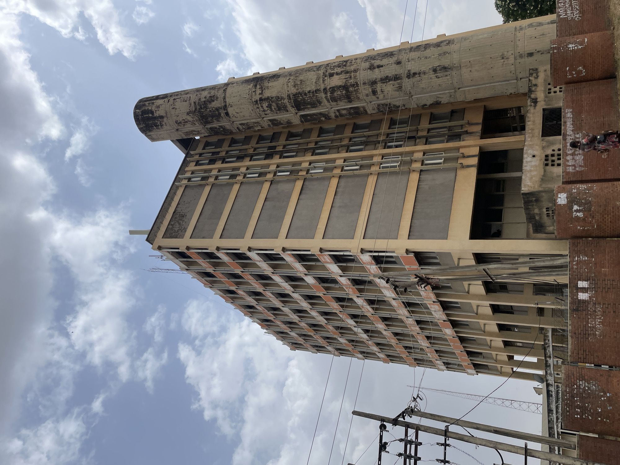 The image shows a tall, rectangular, multi-story building in Jos, Nigeria. The building has a large cylindrical structure on top of it. The building appears to be made of concrete and has a repetitive pattern of rectangular windows or openings. Some windows are covered with gray panels. The building is in a state of disrepair, with visible signs of aging and weathering.
In the foreground, there are power lines and utility poles. To the right of the building, there is a red brick wall with graffiti. A person is standing by the brick wall.
The sky is visible in the background, with scattered clouds.
