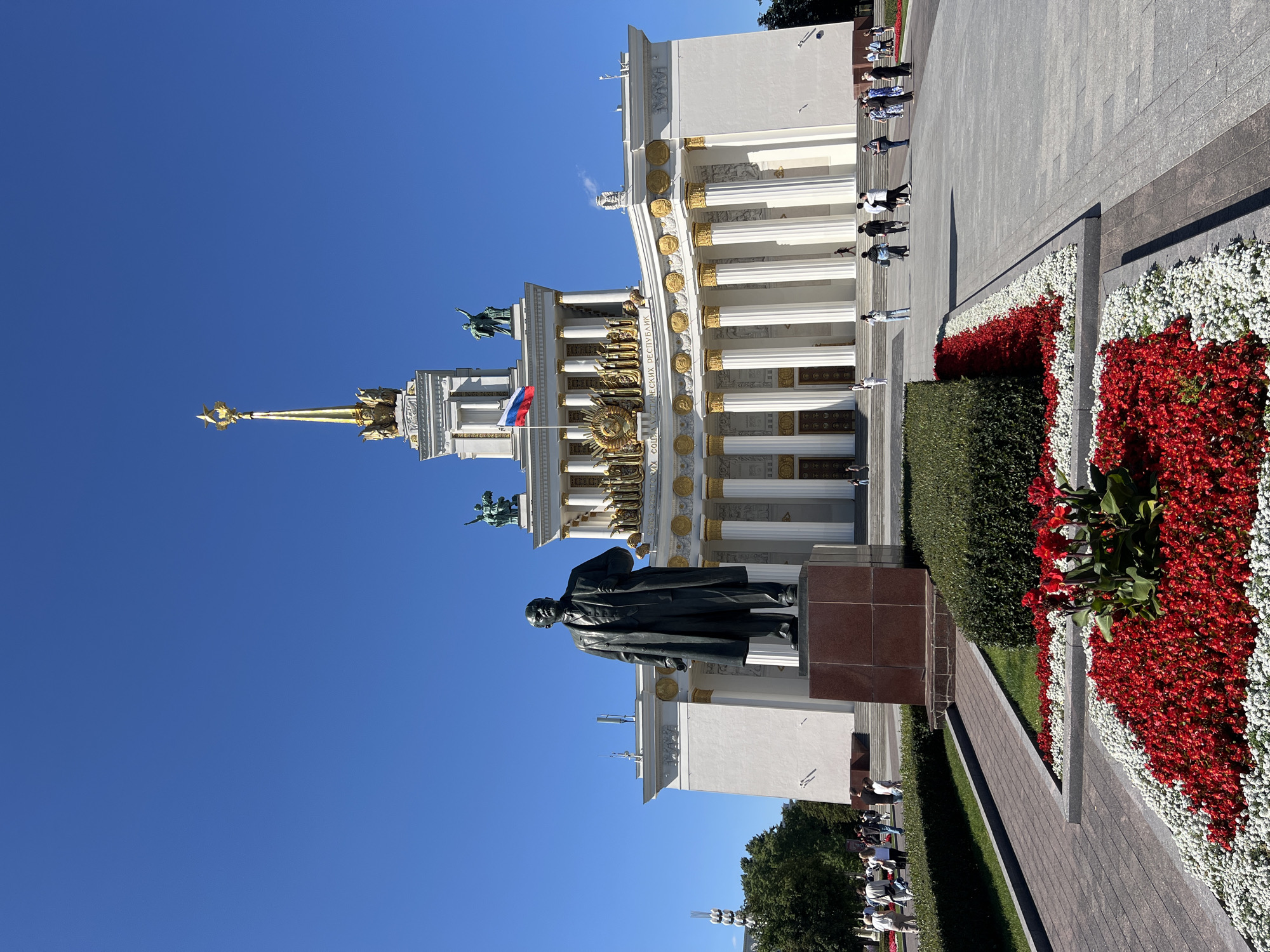 The image depicts the National Exhibition of Economic Achievements (VDNKh) in Moscow, Russia, on a clear day. In the foreground, a statue of Vladimir Lenin stands on a red granite pedestal. The statue faces a grand building with a white facade, golden columns, and ornate architectural details. A Russian flag flies from a flagpole atop the building, which is topped with a golden star. People are visible walking around the plaza and in front of the building. A well-maintained flowerbed with red and white flowers is situated in front of the statue, along with manicured hedges. The sky is a vibrant blue, and trees are visible to the left of the building.