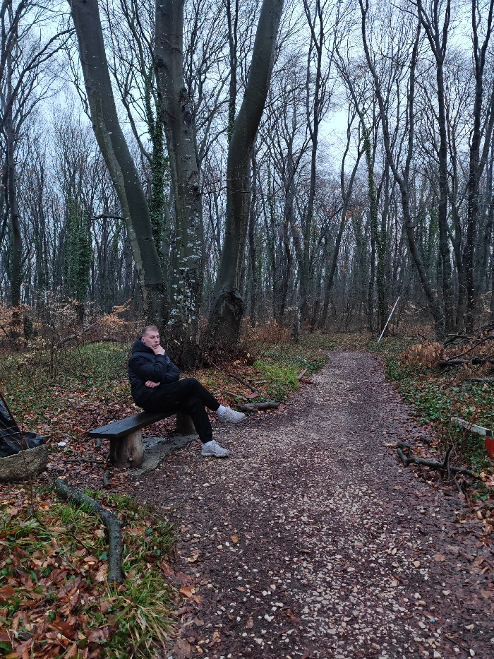 The image captures a young adult male seated on a rustic wooden bench within a forest area in Shumen, Bulgaria. The male is dressed in a black hooded puffer jacket, black pants, and light grey athletic shoes. He is positioned with his legs crossed at the ankles, his right arm across his chest, and his left hand raised near his chin, suggesting a contemplative posture, as he looks towards the right. The environment consists of a dense deciduous forest with numerous tall trees, most bare of leaves, indicating late autumn or winter. Some tree trunks are partially covered with green ivy. The ground is a forest floor covered extensively with brown and orange fallen leaves, interspersed with patches of green vegetation. A winding, gravelly dirt path, appearing damp, stretches from the foreground into the background. To the left of the bench, a metal container or makeshift fire pit is visible on the ground. Further along the path to the right, a thin white pole stands upright, and a red and white striped barrier is partially visible in the bottom right corner of the frame. The sky is overcast, providing soft, diffused lighting.